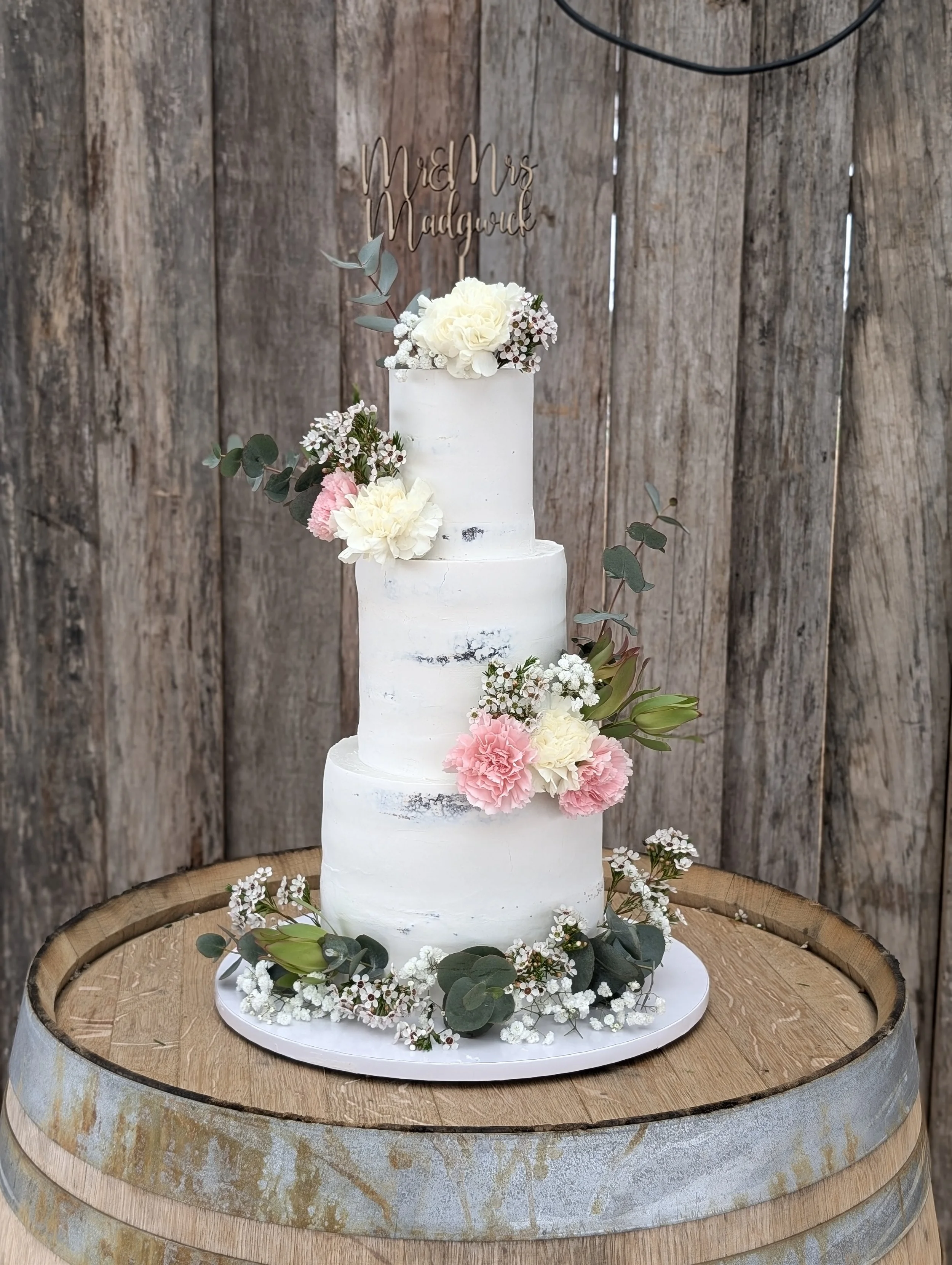 A three-tier white wedding cake with a semi-naked frosting style, decorated with white, pink, and cream flowers, greenery, and a gold cake topper that reads 'Mr & Mrs Madgwick,' placed on a wooden barrel against a rustic wooden backdrop.