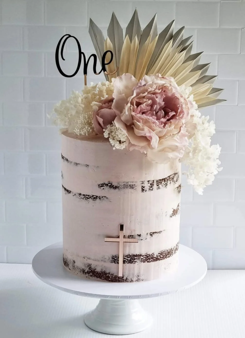 A white cake with a marbled black and white finish, decorated with large pink and cream flowers, white hydrangeas, and a bouquet of gold-colored leaves on top. A black cursive decoration that reads 'One' and a small wooden cross are also on the cake.