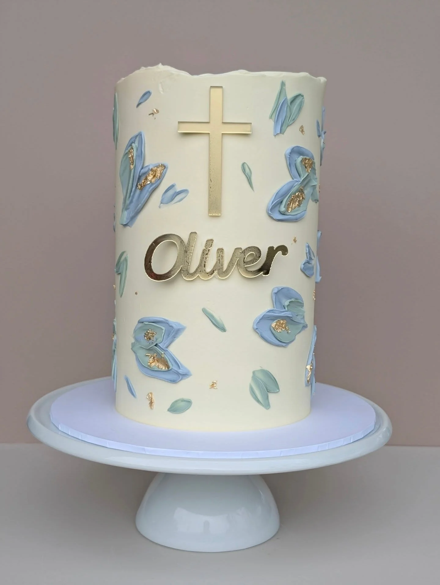 A white cake decorated with blue and gold floral designs, featuring a gold cross at the top, and the name 'Oliver' in gold lettering. The cake is on a white cake stand.
