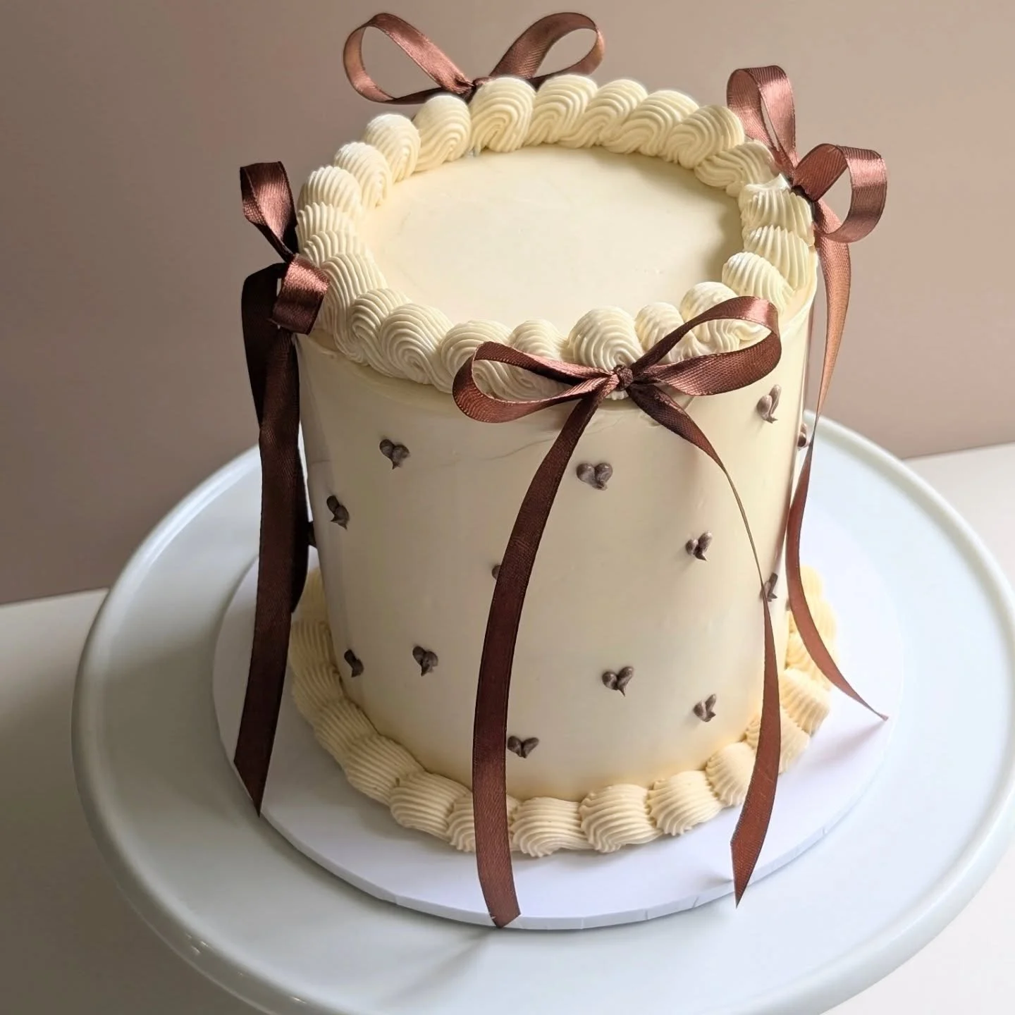 A tall white celebration cake decorated with brown ribbons and small dark brown heart-shaped accents, topped with a border of piped cream and sitting on a white cake board.
