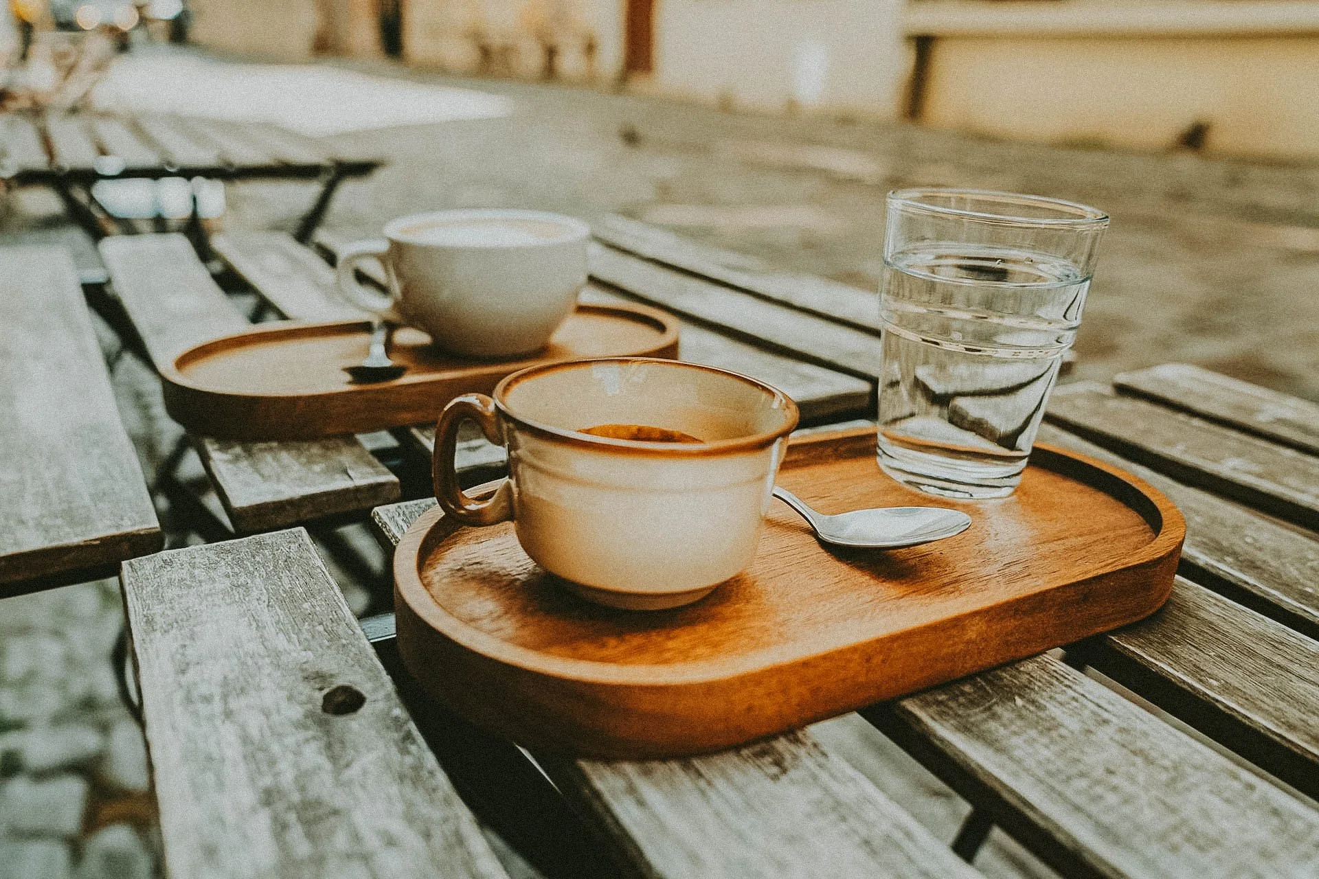 A wooden tray with a cup of coffee, a glass of water, and a spoon on a wooden table outdoor setting.