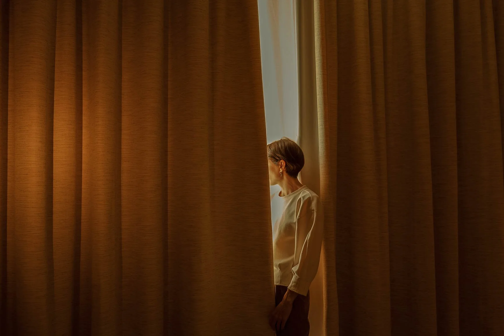 A woman peeking from behind a beige curtain in a softly lit room.