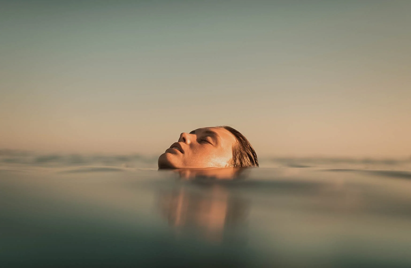 Close-up of a woman swimming with only her head emerging from the water, eyes closed, during sunset or sunrise.