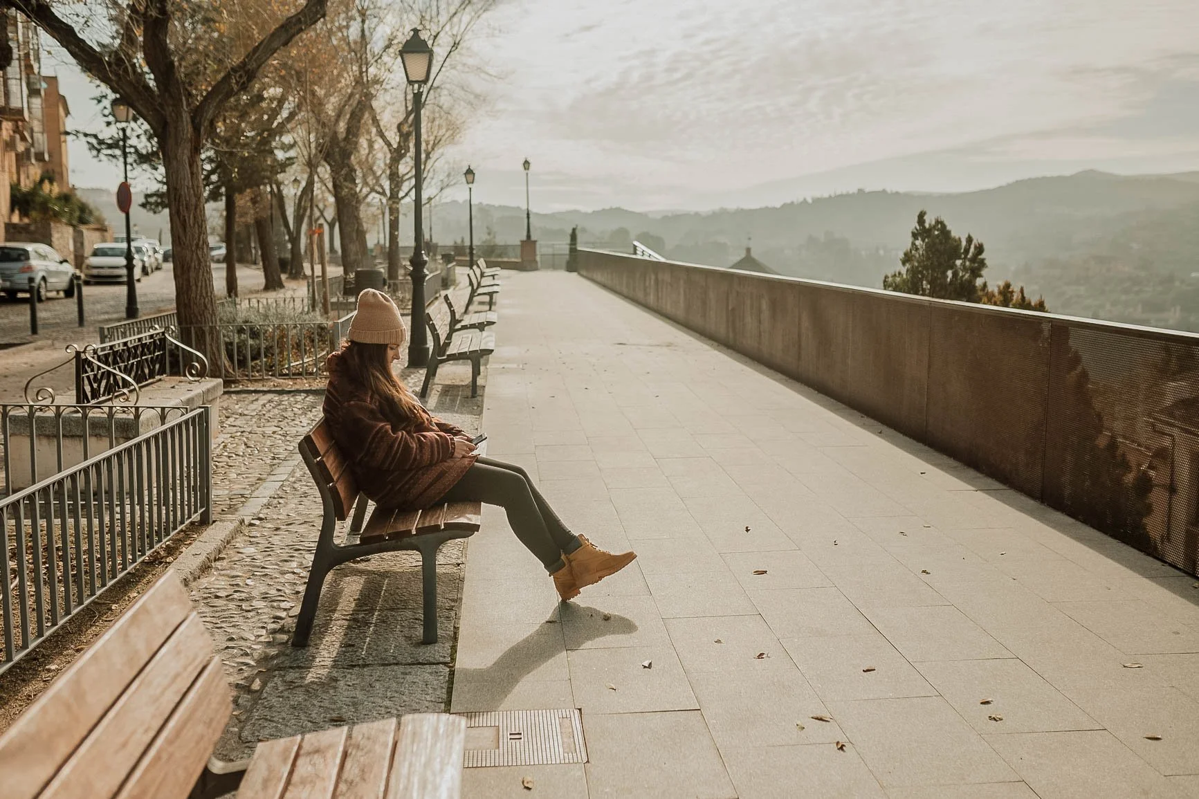 A woman sitting on a park bench using her phone, wearing a beige beanie, brown jacket, black pants, and tan boots, with a scenic view of mountains in the background and a walkway lined with lamps and additional benches.