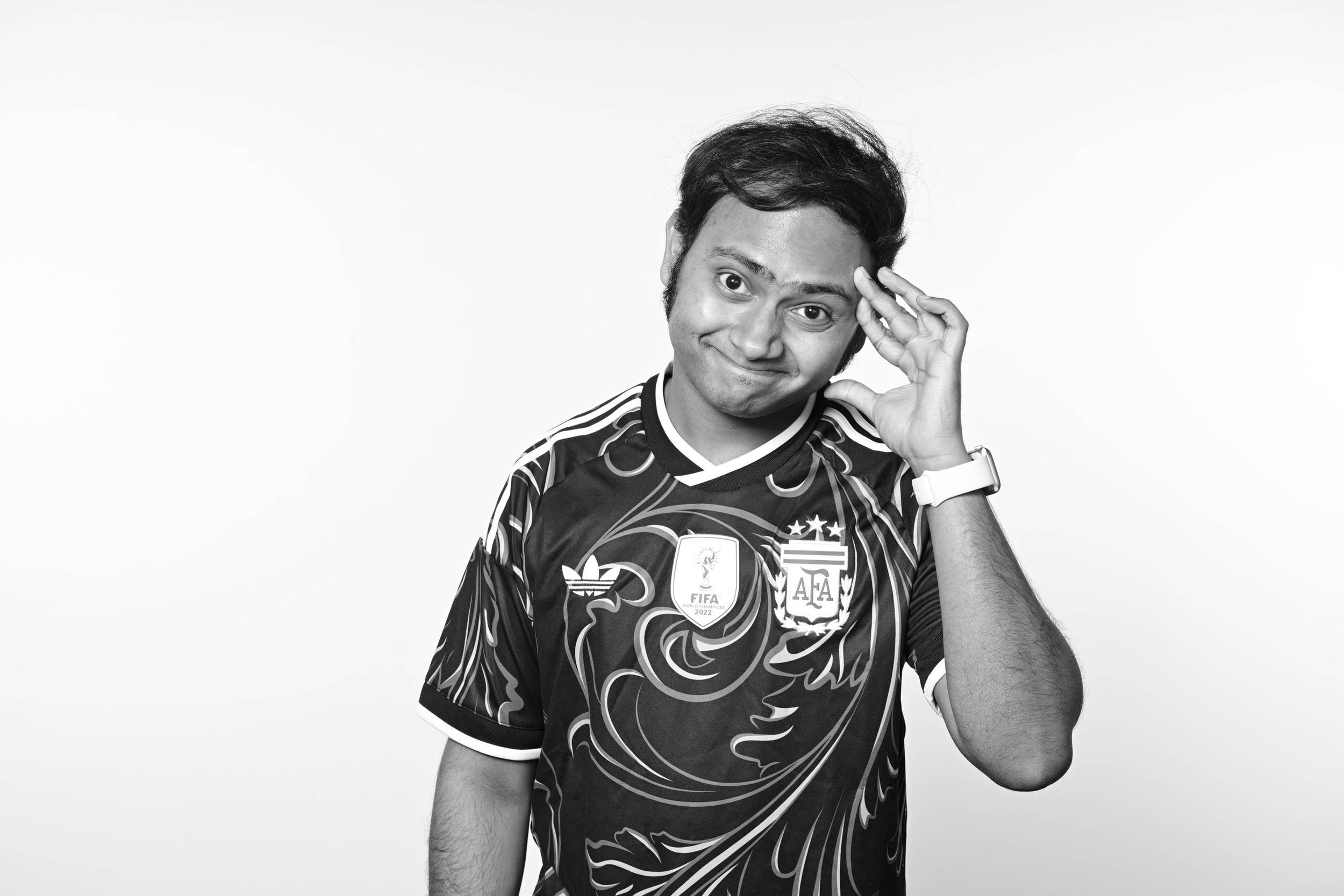 Black and white photo of a young man with dark hair wearing an Argentina soccer jersey with various logos, holding his hand to his head and smiling.