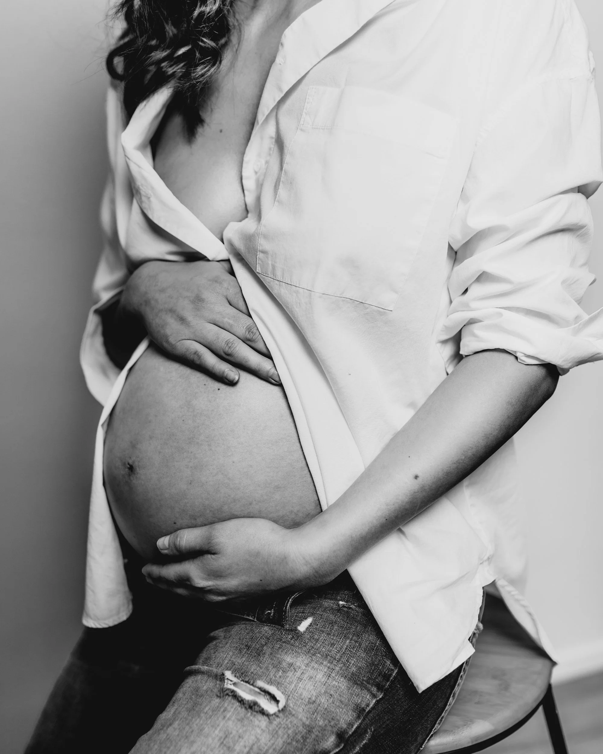 A pregnant woman in a loosened white shirt and ripped jeans, sitting and cradling her pregnant belly with both hands.