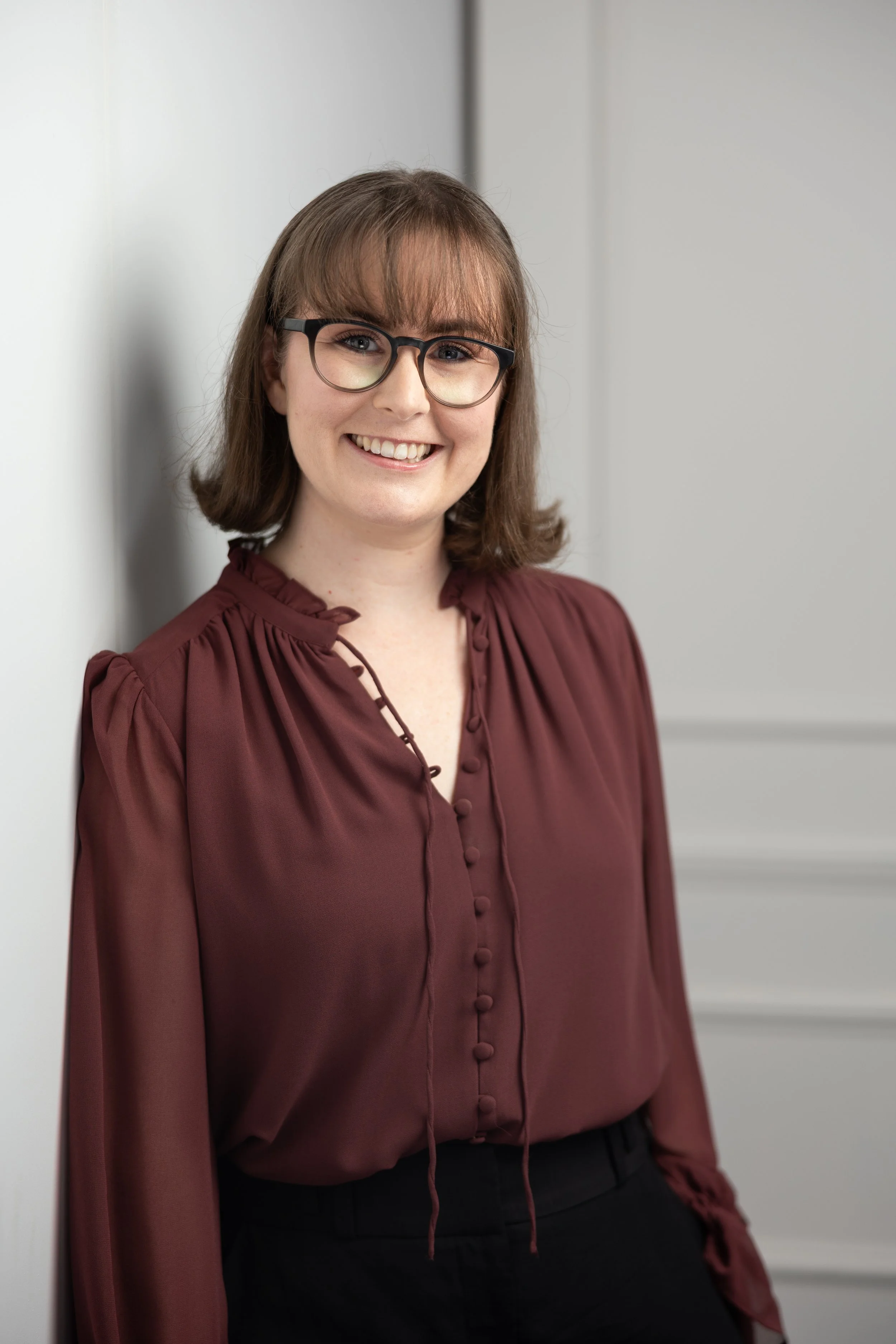 A woman with short brown hair and glasses smiling while leaning against a wall