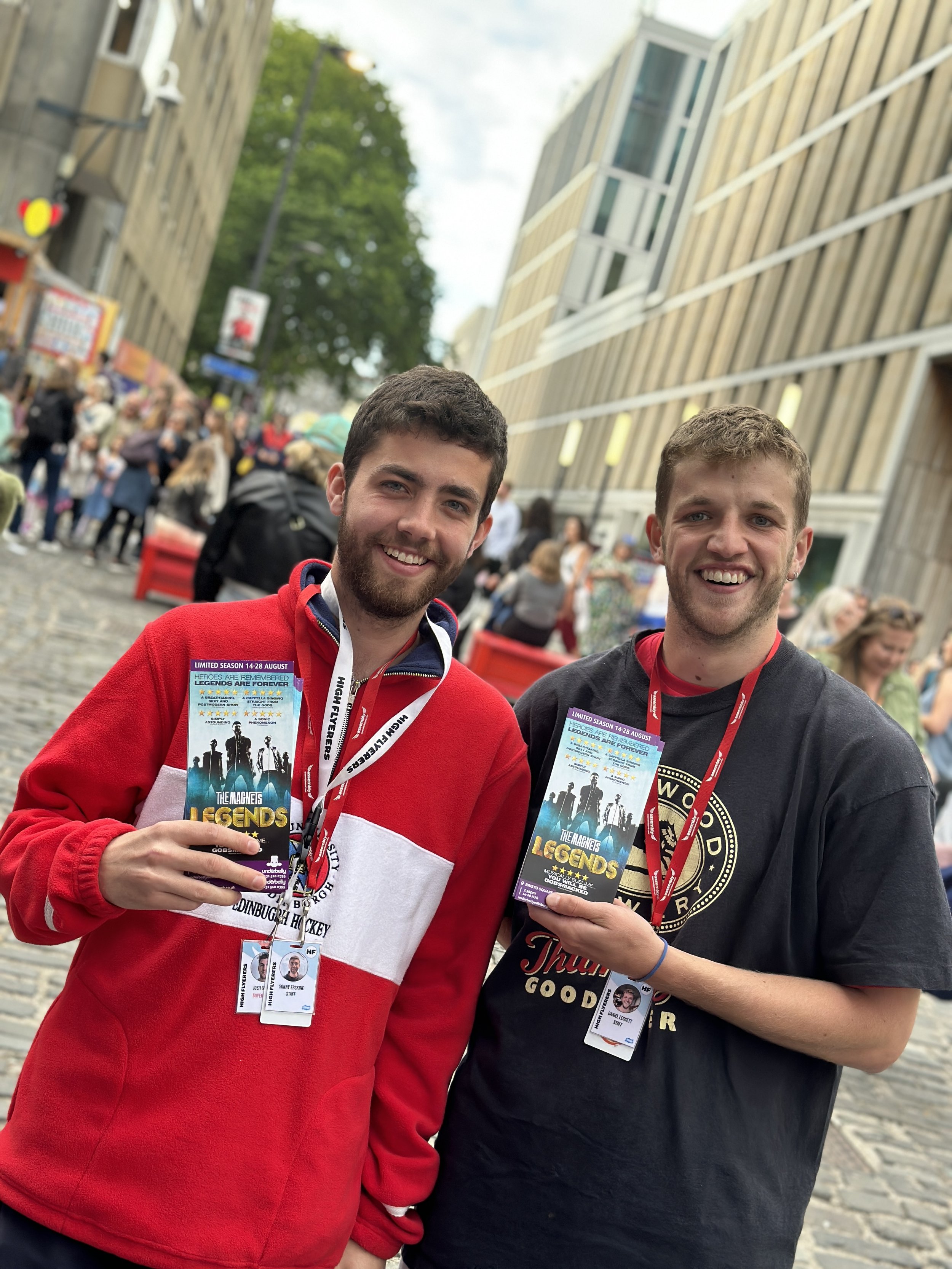 Two members of our team smiling and holding flyers for a show called 'Legends' on a busy Edinburgh city street, during the Edinburgh Fringe Festival.