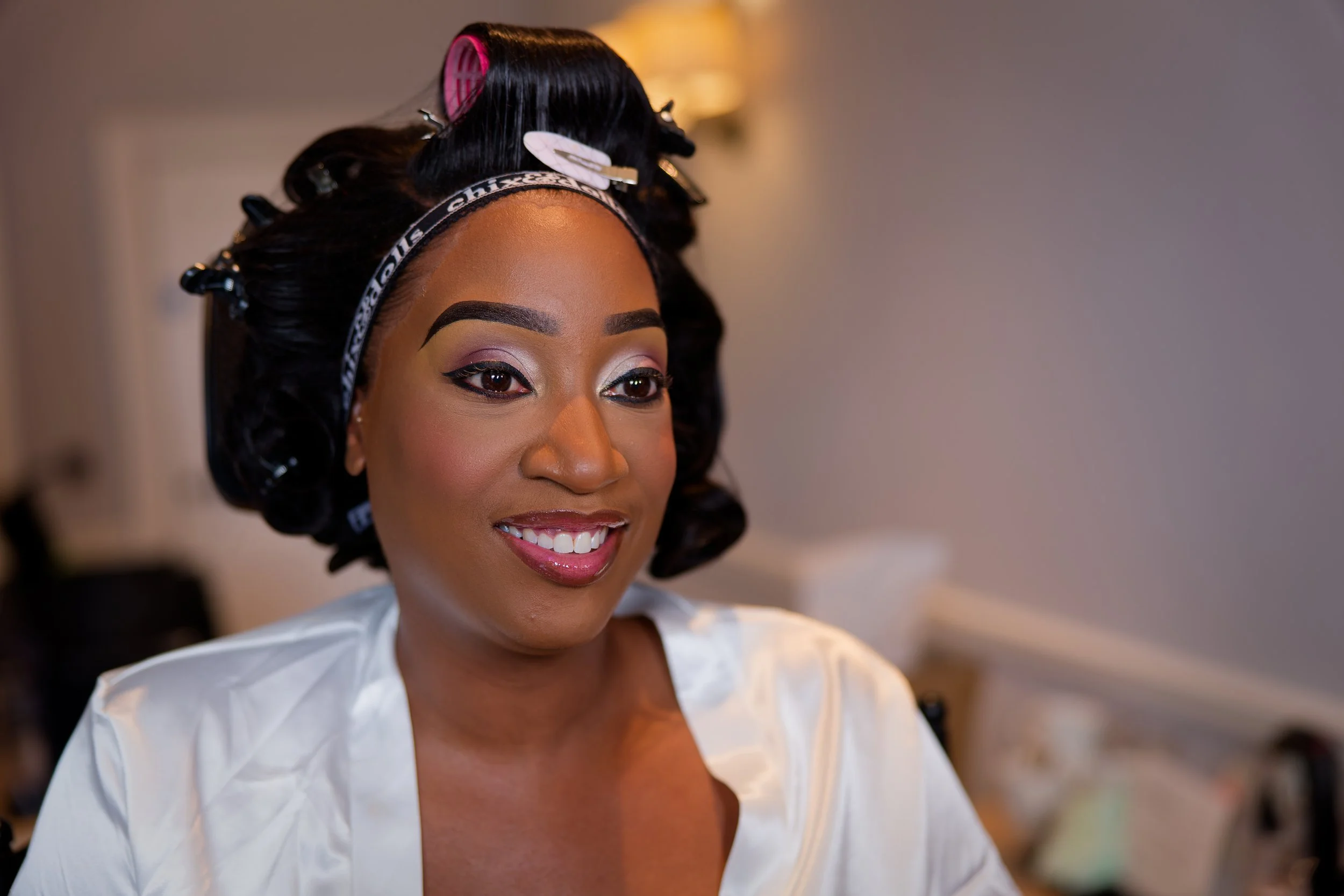 A woman with styled black hair and makeup, wearing a white satin robe, smiling, with hair rollers and a black and white headband.