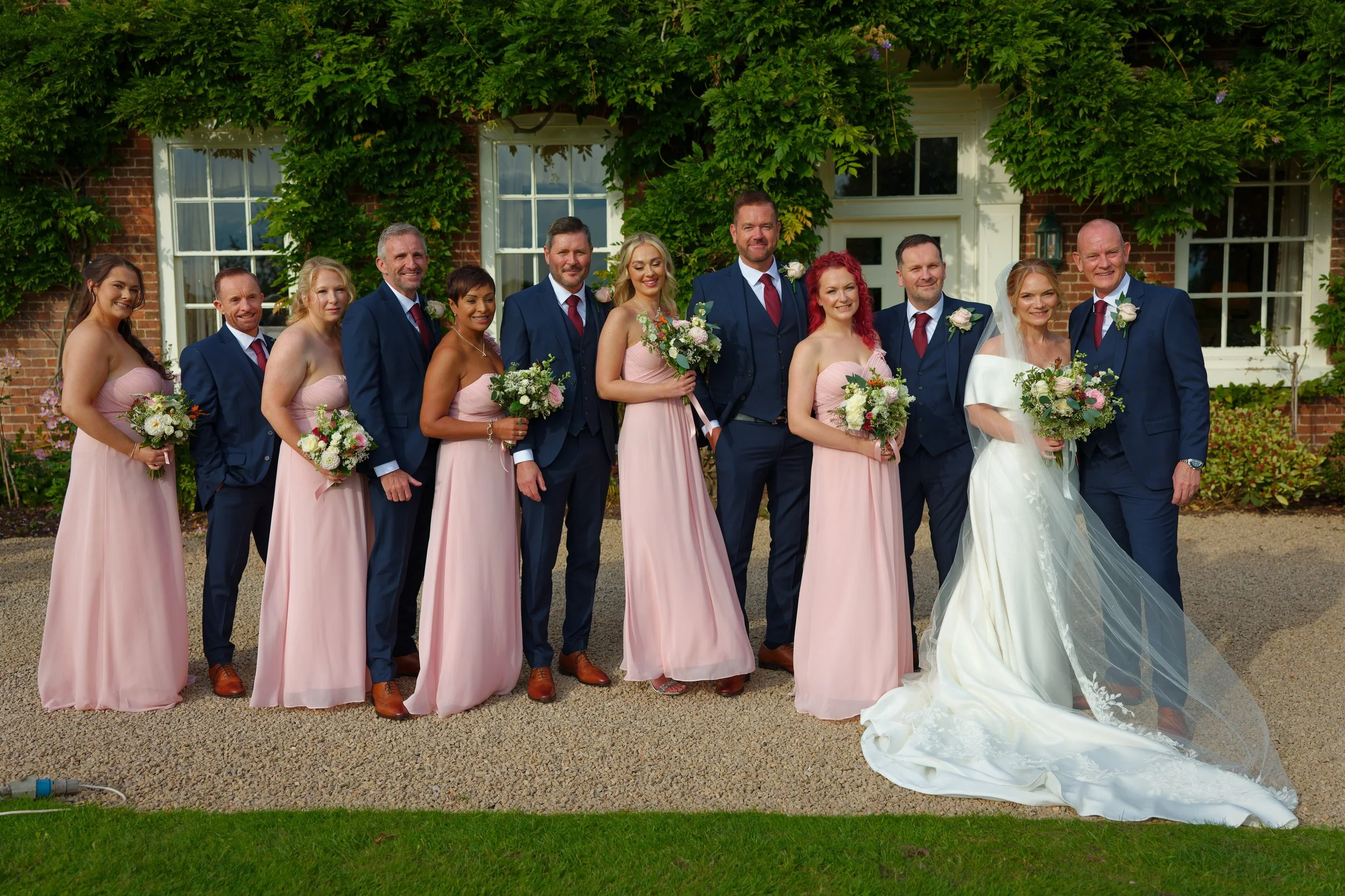 Group of people in wedding attire standing outdoors in front of a brick building with greenery, including a bride in a white gown with a long veil and veiled train, and bridesmaids in blush pink dresses holding bouquets, along with groomsmen in navy 