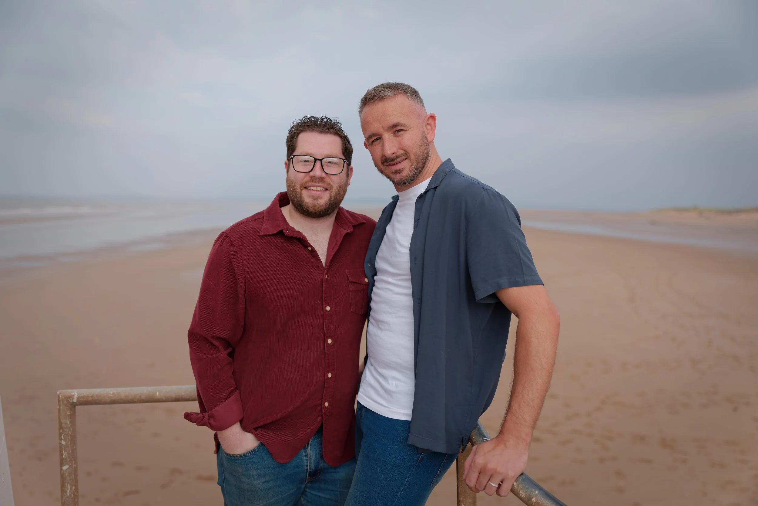 Two men standing on a beach with sand and ocean in the background, posing together for a photo.