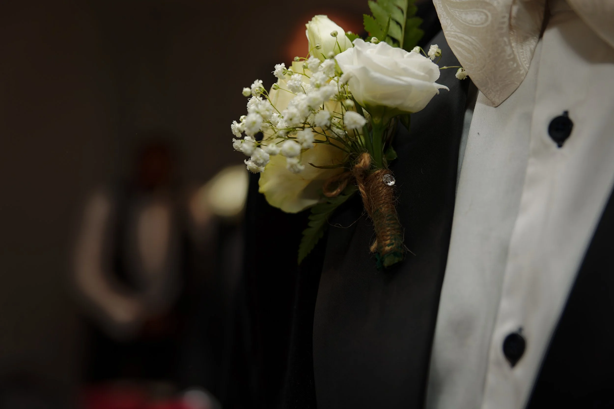 Close-up of a white boutonniere with small white flowers and green leaves pinned to a black suit jacket at a formal event.