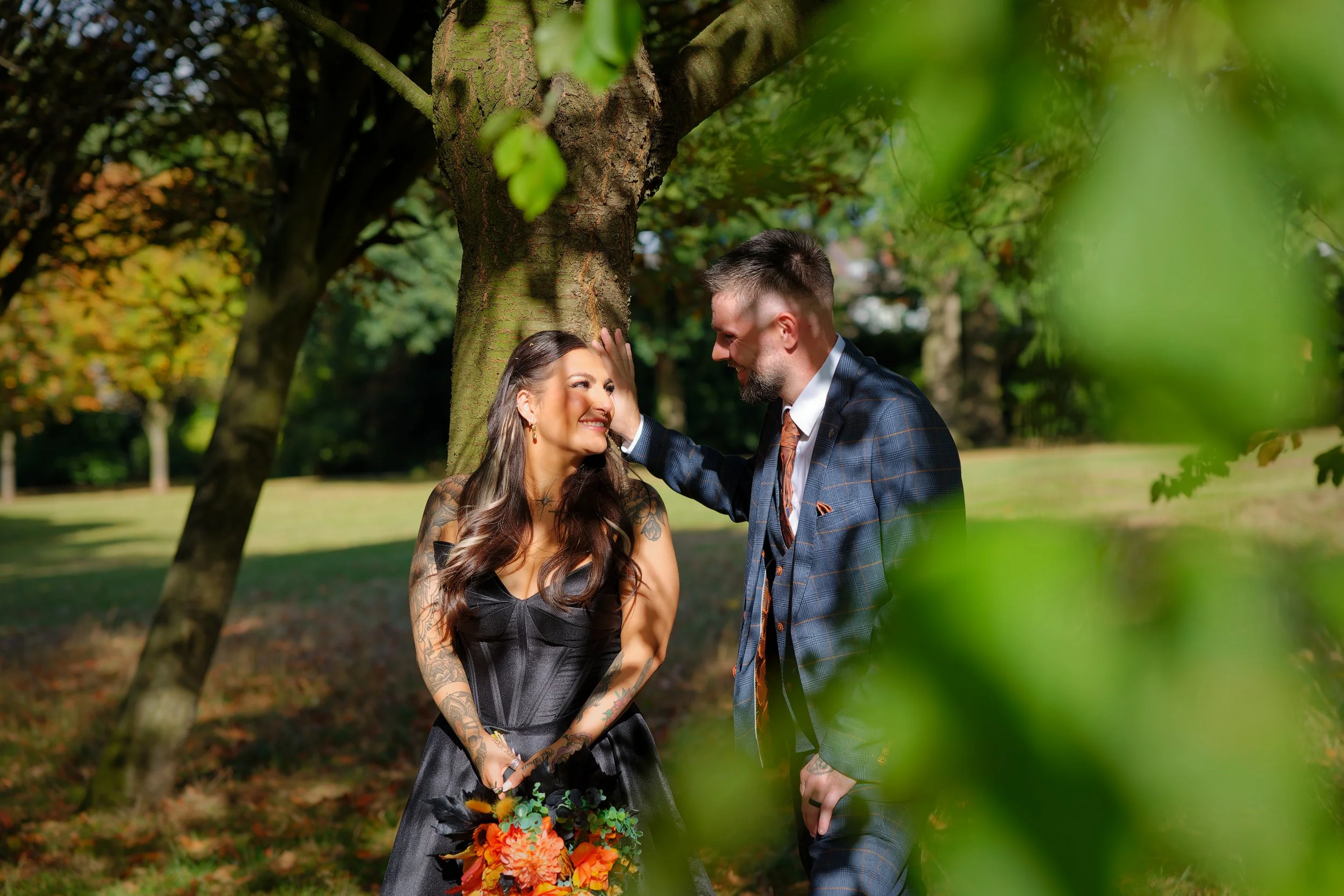 A couple standing outdoors near a tree, gazing at each other and smiling, with woman holding a bouquet of flowers.