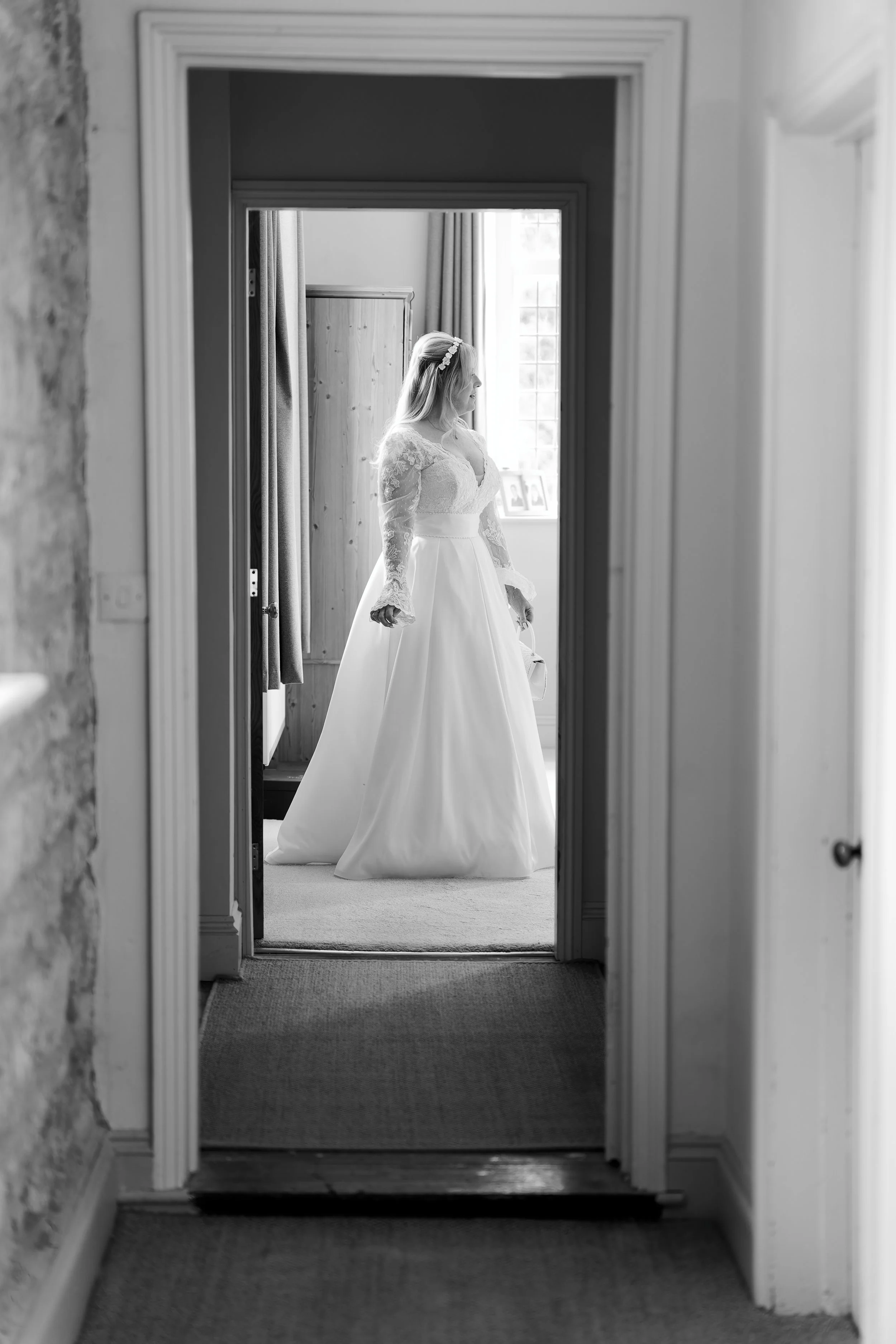 A bride in a wedding dress standing in a room, viewed through a doorframe, with light coming from a window behind her.