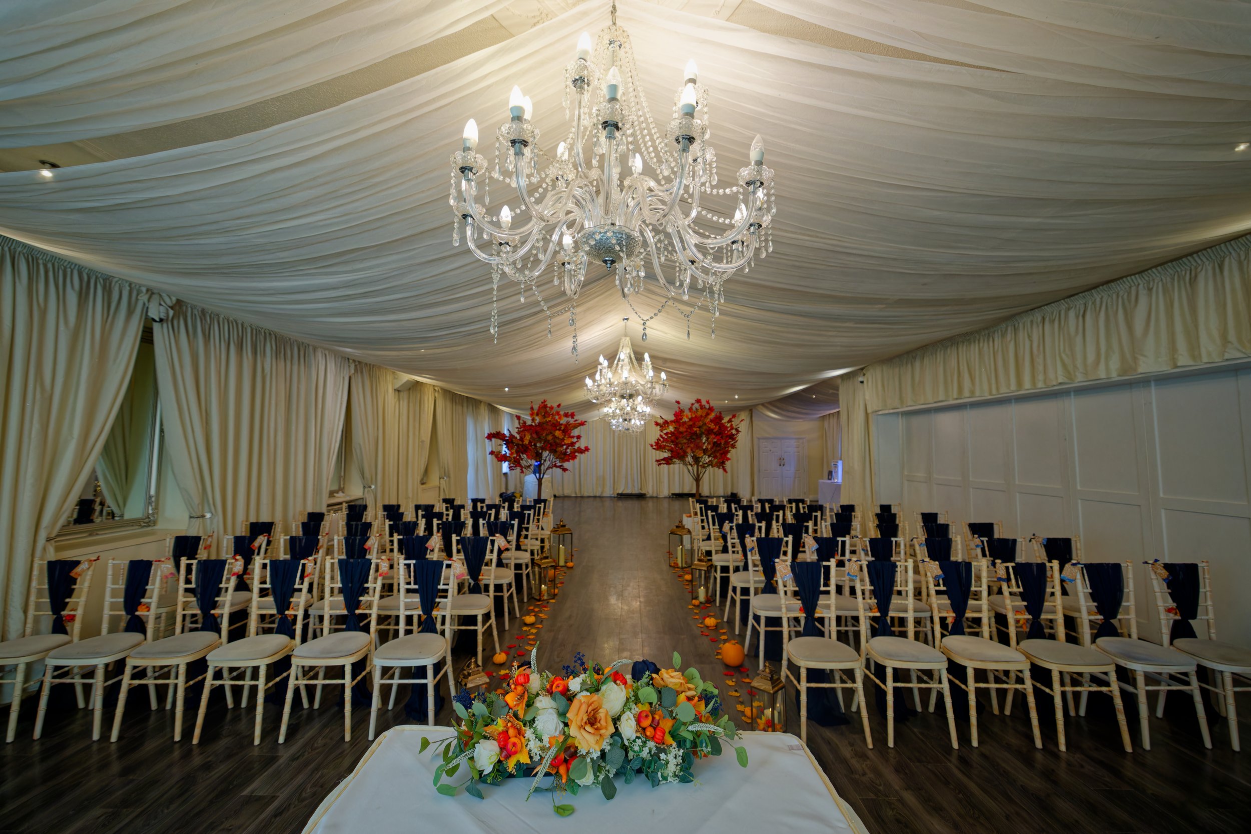 Elegant wedding ceremony setup with white chairs draped with navy sashes, a floral aisle runner with orange and yellow flowers, and red trees at the altar. Chandeliers hang from the draped ceiling in a decorated, cream-colored tent.