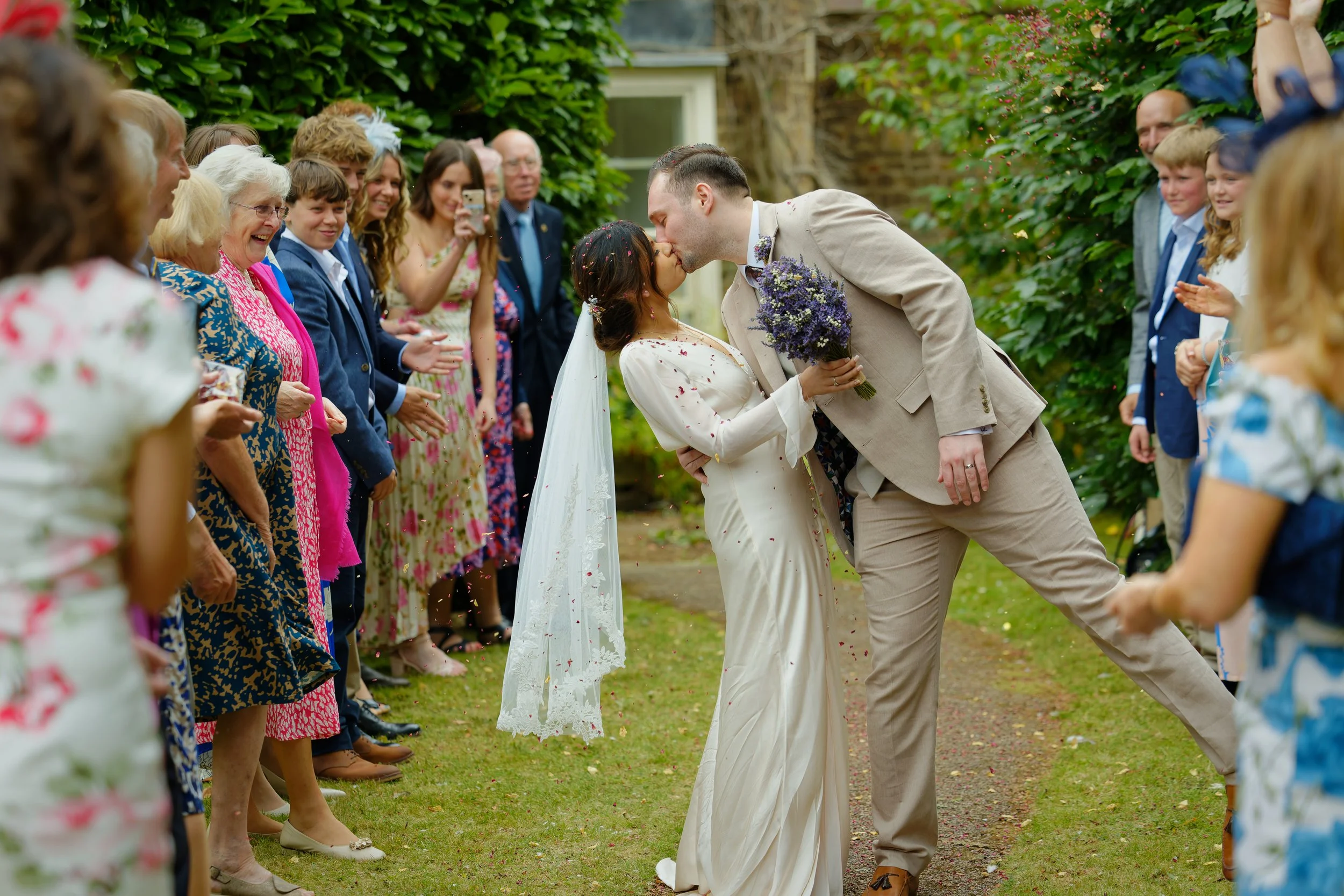 A newlywed couple sharing a kiss outdoors, surrounded by wedding guests, with the groom holding a bouquet and guests clapping and celebrating.