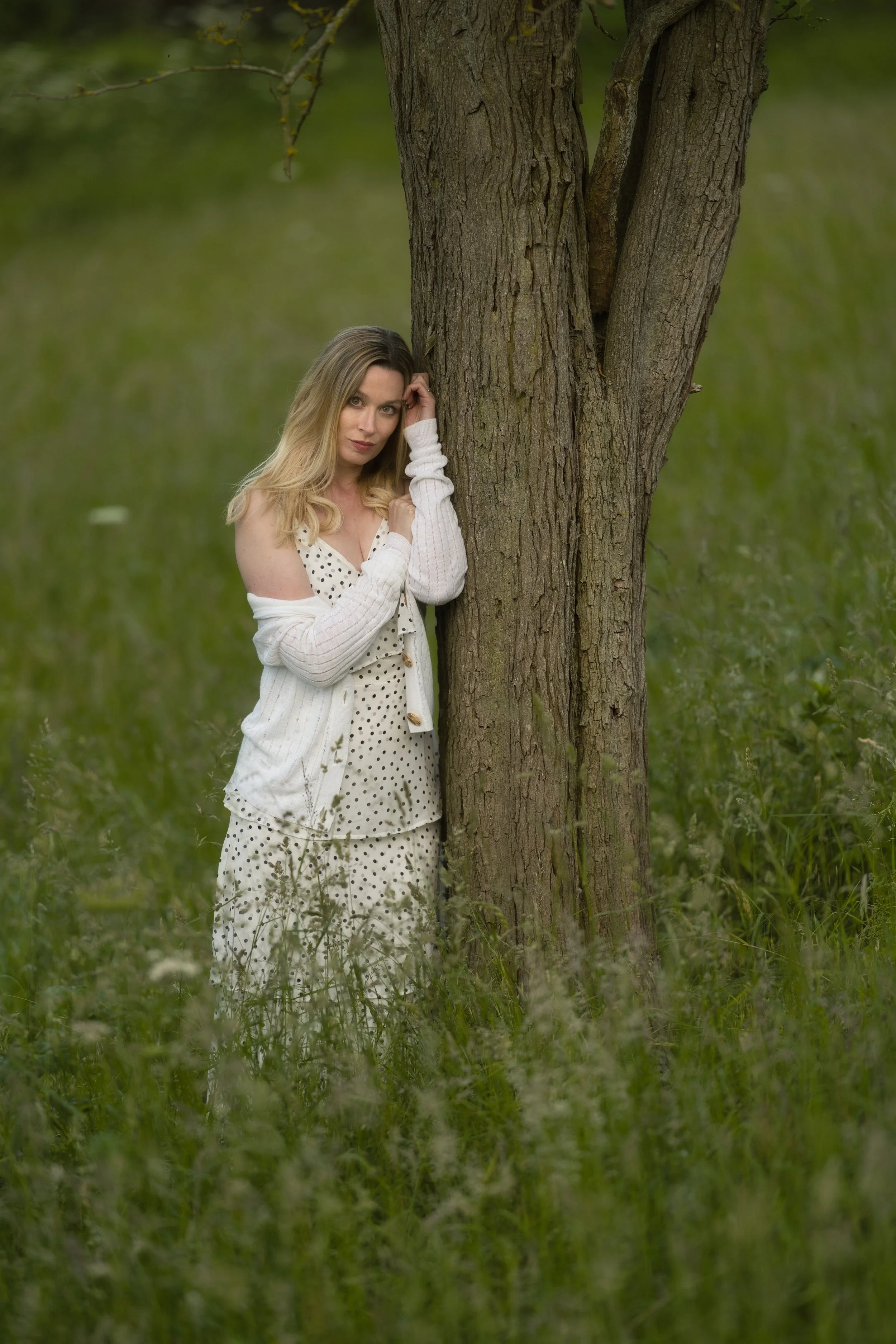A woman with blonde hair in a polka dot dress and cardigan standing beside a large tree in a grassy outdoor area.