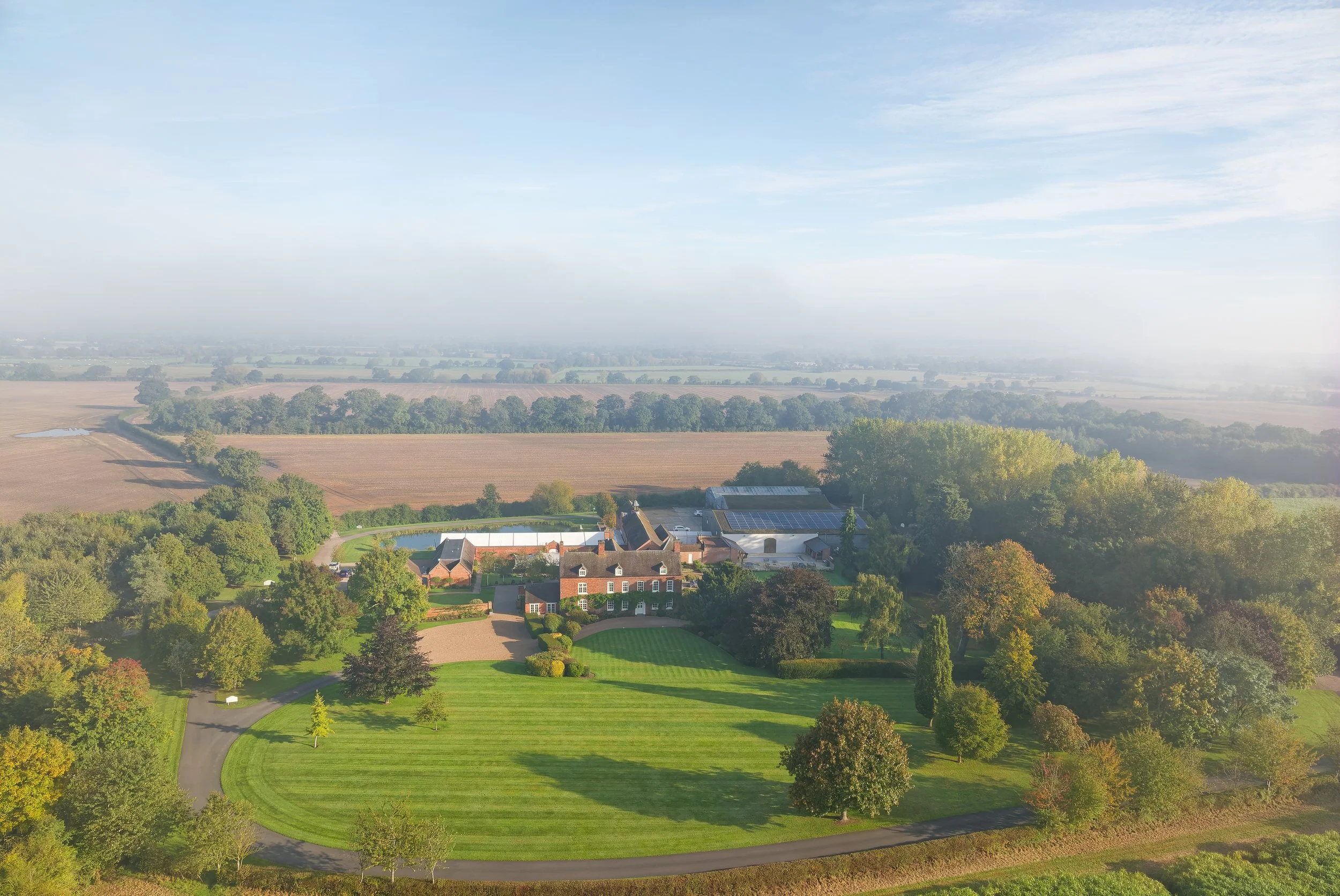 Aerial view of a large estate with a mansion, garden, trees, and surrounding farmland on a sunny day.