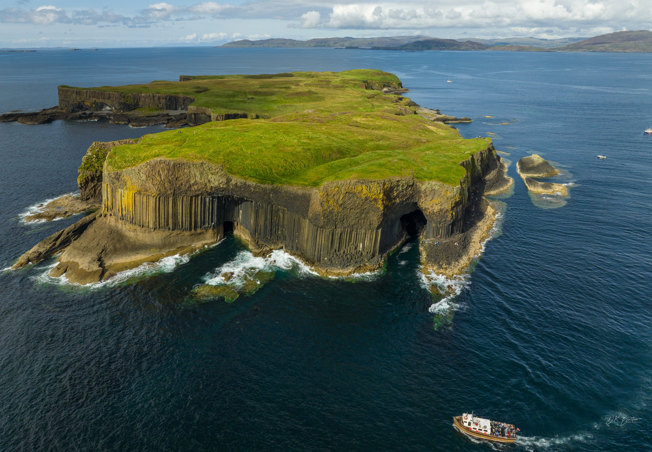 Aerial view of a green island with cliffs and caves, surrounded by ocean, with boats near the shore and distant landmass in the background.