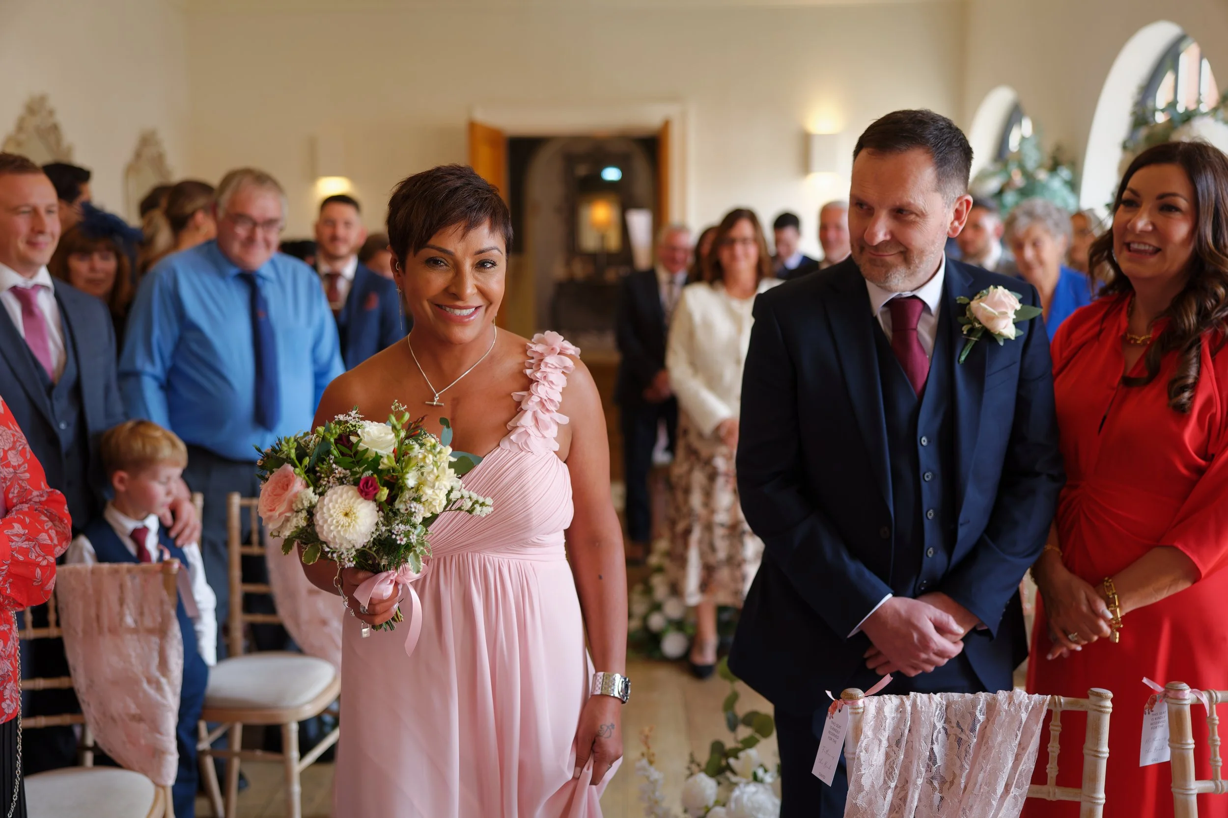 A woman in a pink dress holding a bouquet, standing in a decorated room with wedding guests.