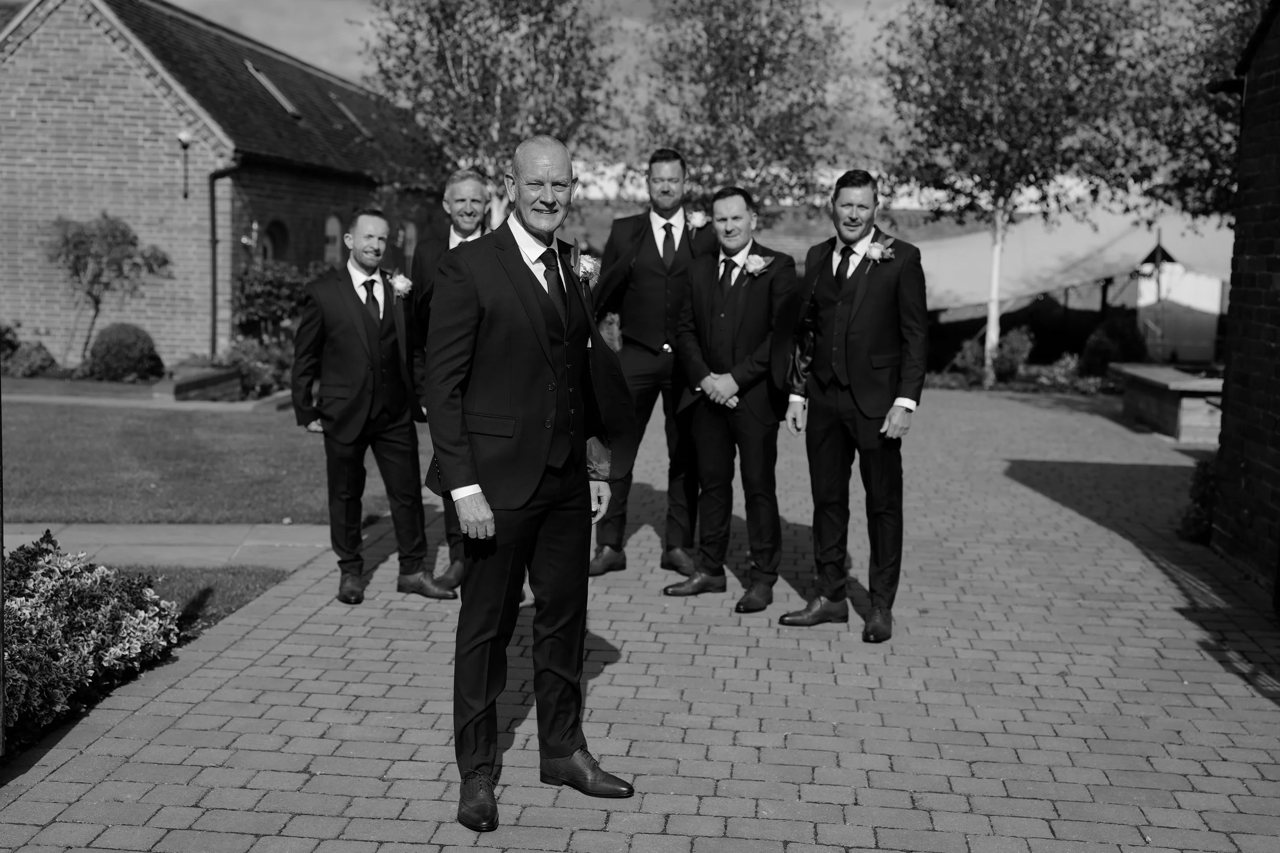 Group of men in suits standing outside on a sunny day, posing for a photo at a wedding or formal event.