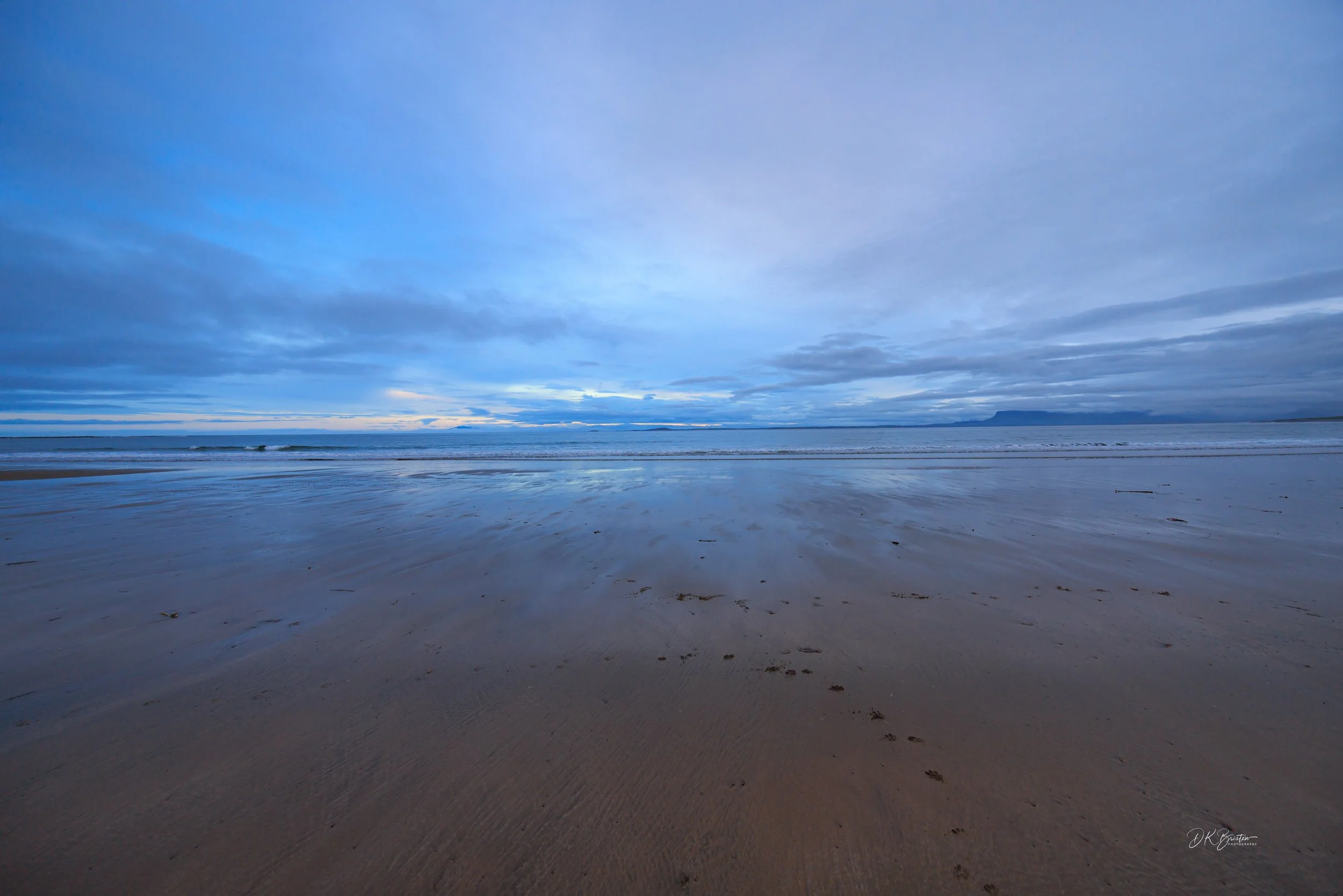 A serene ocean beach at dusk with wet sand reflecting blue sky and scattered clouds on the horizon.