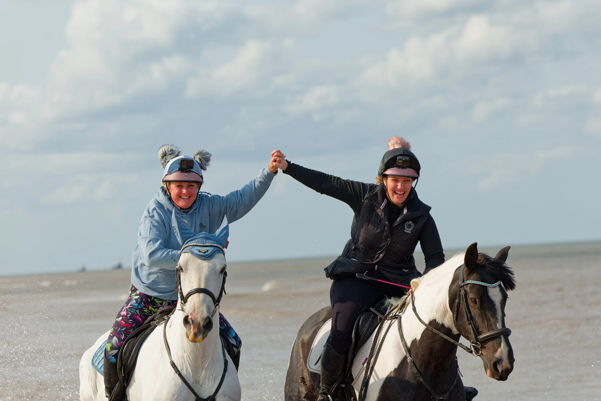 Two women riding horses on the beach, holding hands and smiling, with the ocean and sky in the background.
