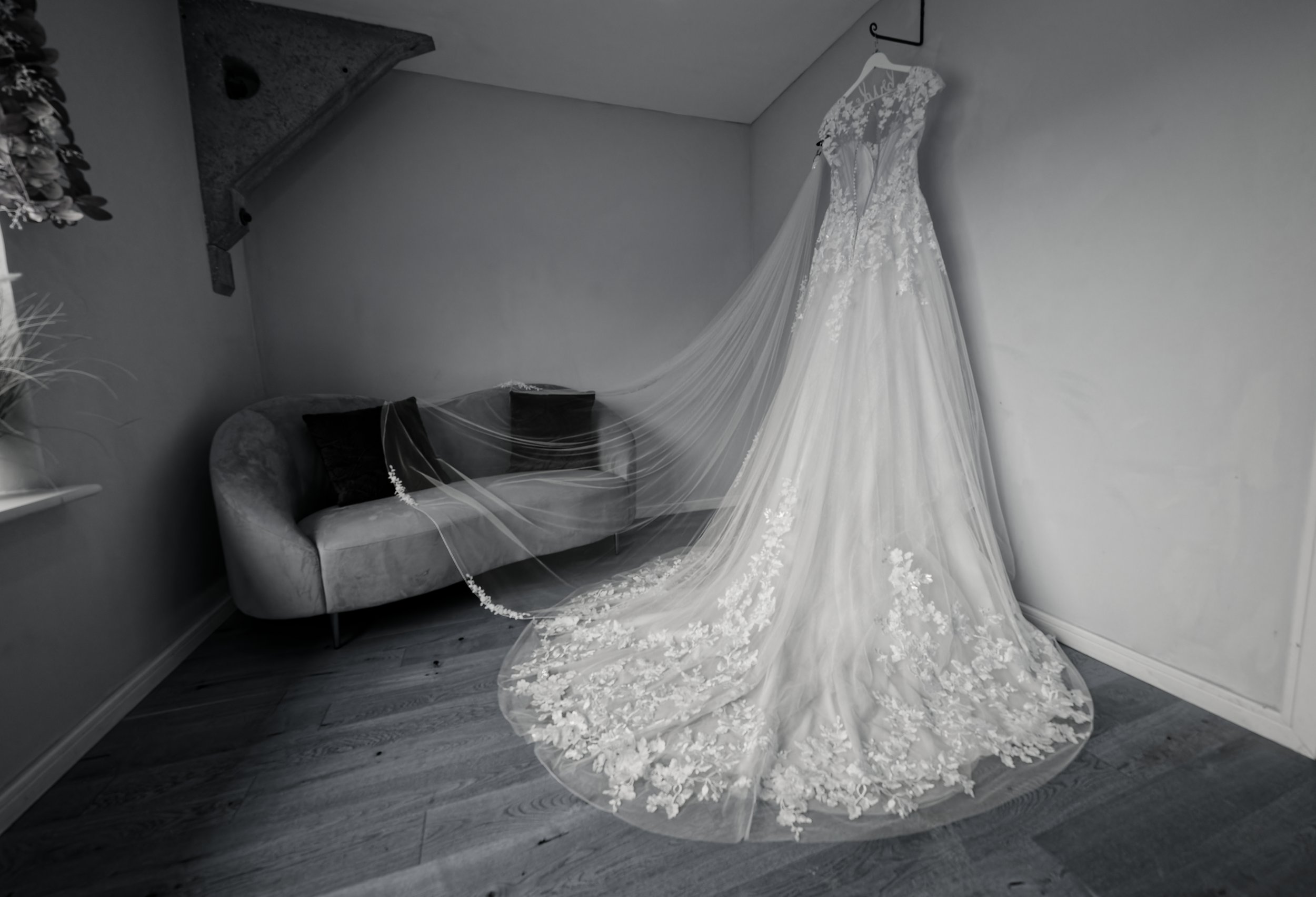 A wedding dress with lace details and a long train hanging on a wall hook in a room. A small velvet sofa with two black pillows is next to it.