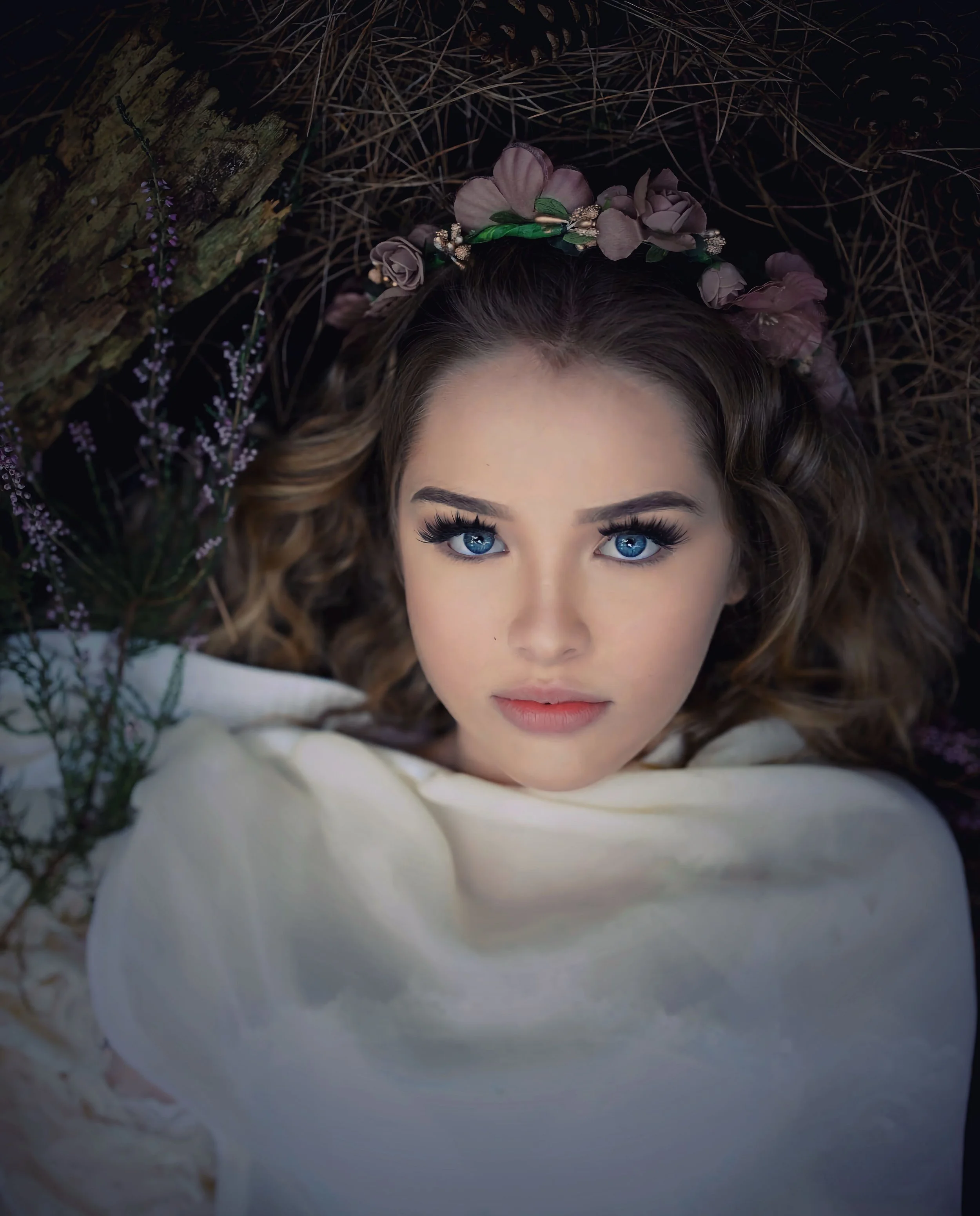 A woman with blue eyes and wavy brown hair lying on the ground, surrounded by pine needles and purple flowers, wearing a floral headband and a white garment.