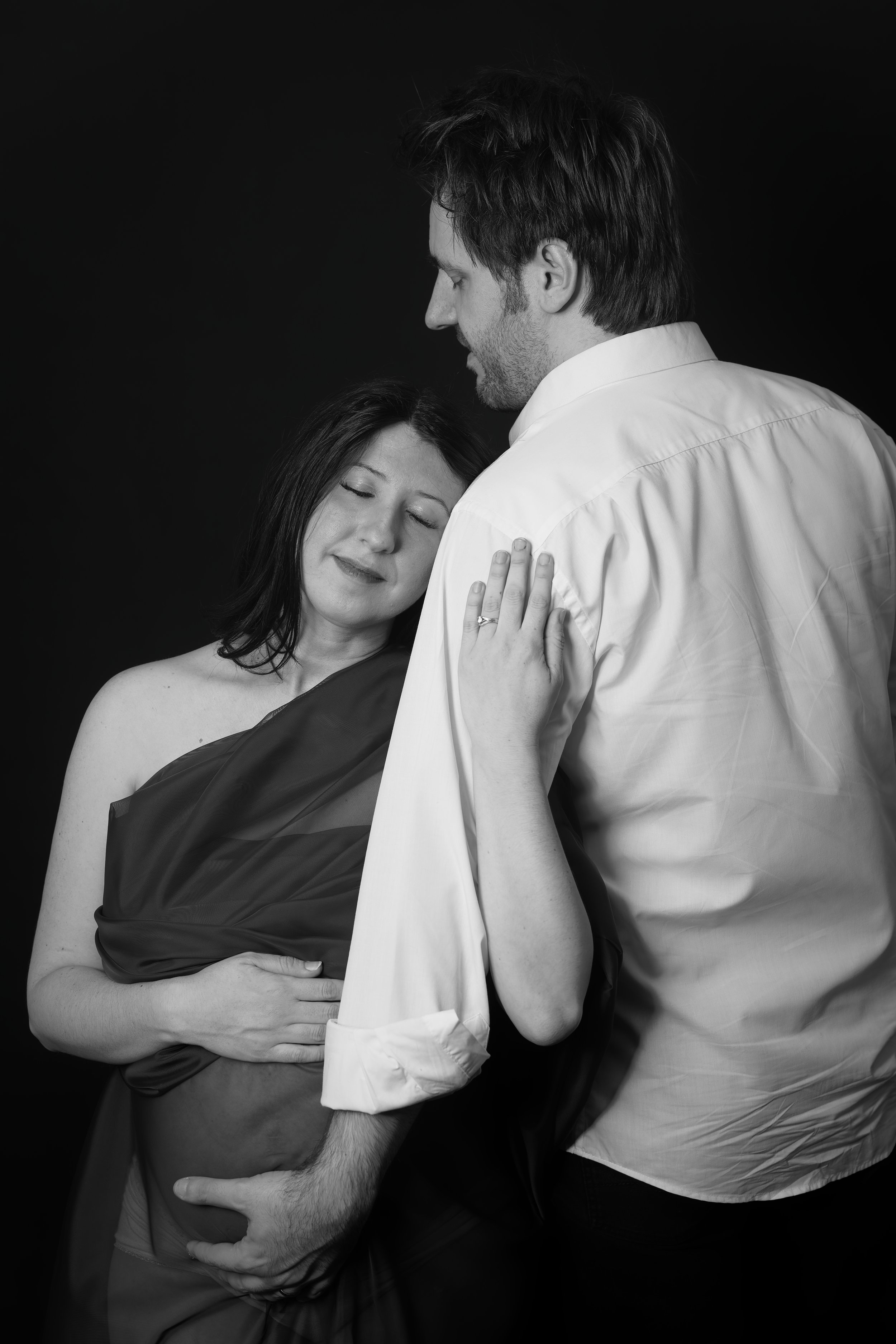A couple, with the woman visibly pregnant, sharing an intimate moment against a dark background. The woman is gently holding her belly with her eyes closed, while the man is embracing her and looking down at her.