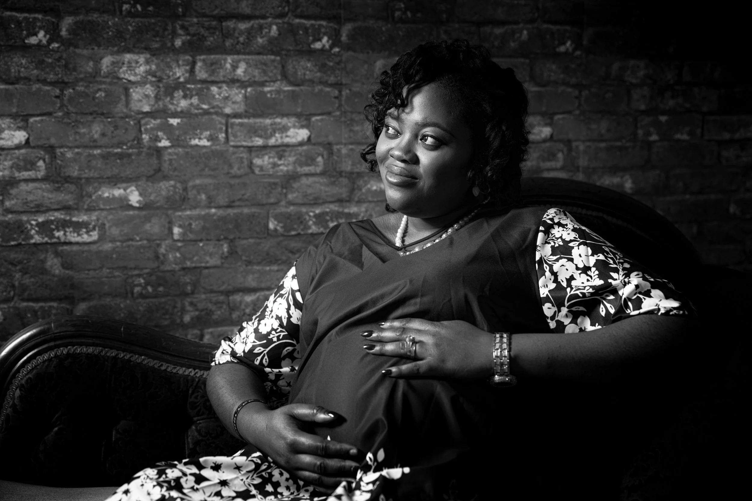 A pregnant woman with dark curly hair sitting on a vintage-style chair against a brick wall, wearing a patterned dress and jewelry, looking thoughtful.