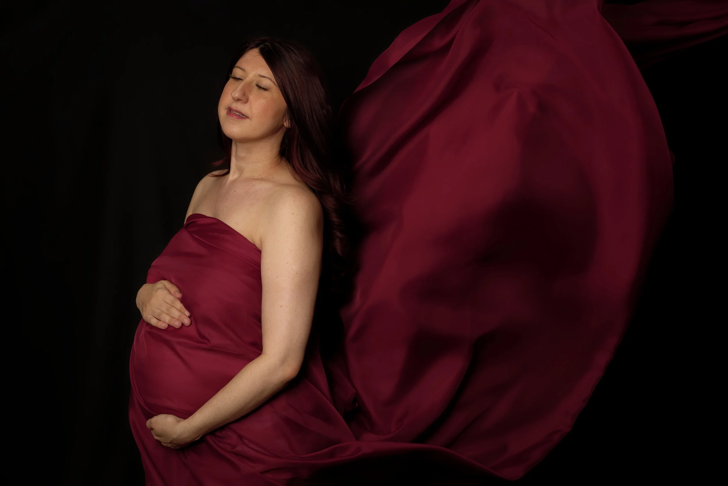Pregnant woman in a maroon dress with her eyes closed, standing against a black background, with flowing fabric extending from her dress.
