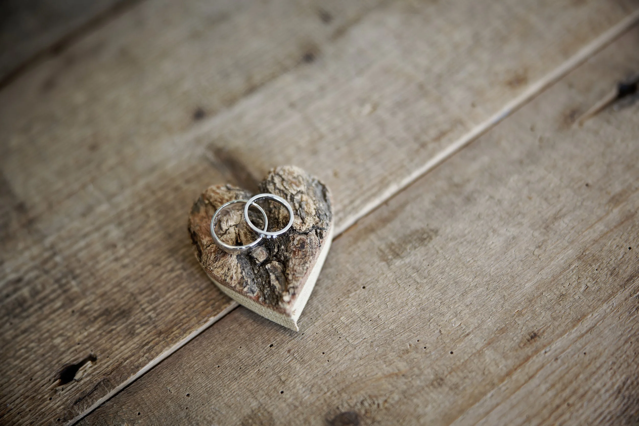 Two silver rings placed on a piece of wood shaped like a heart, on a rustic wooden surface.