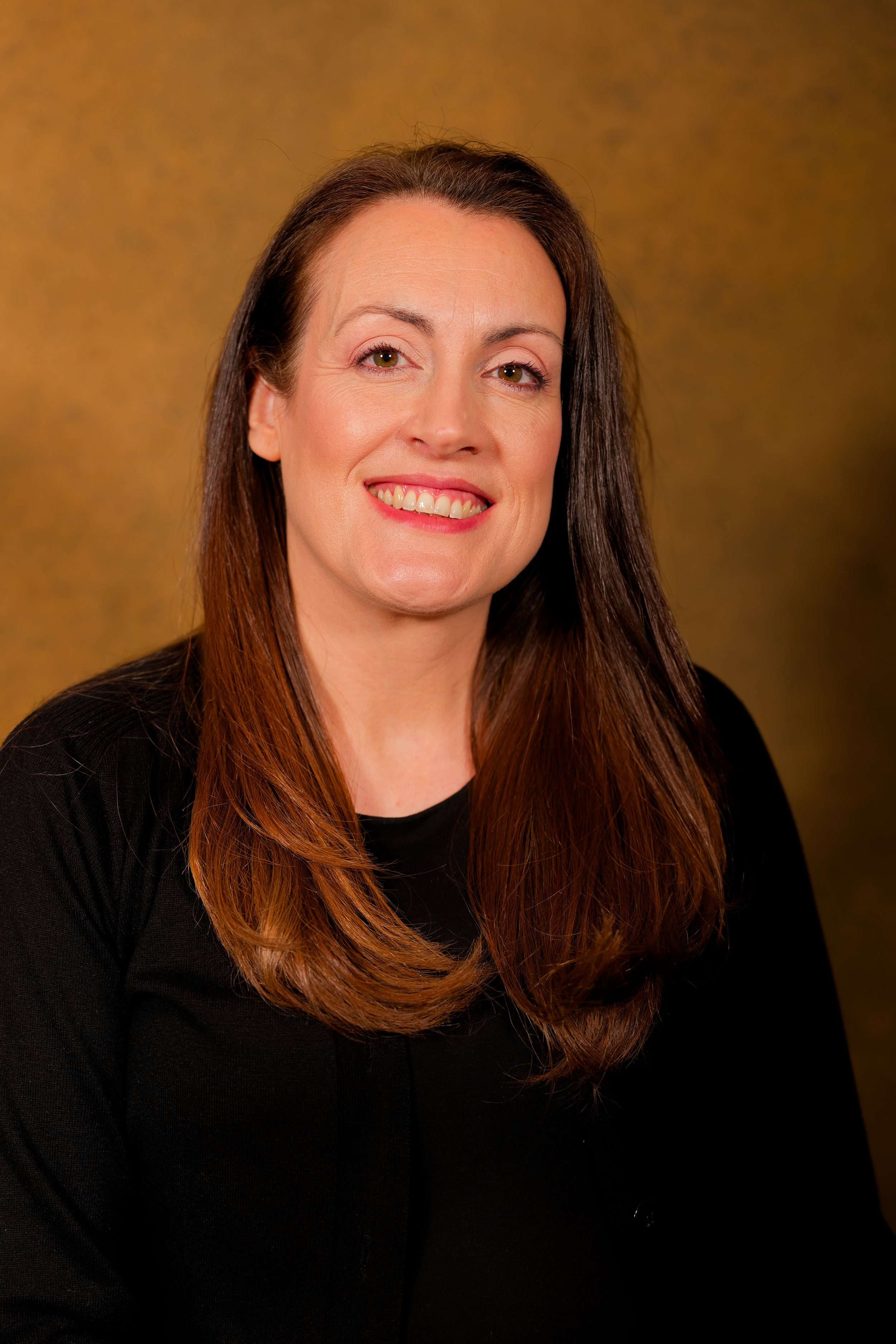 A woman with long brown hair and fair skin smiling, wearing a black top, against a golden-brown background.
