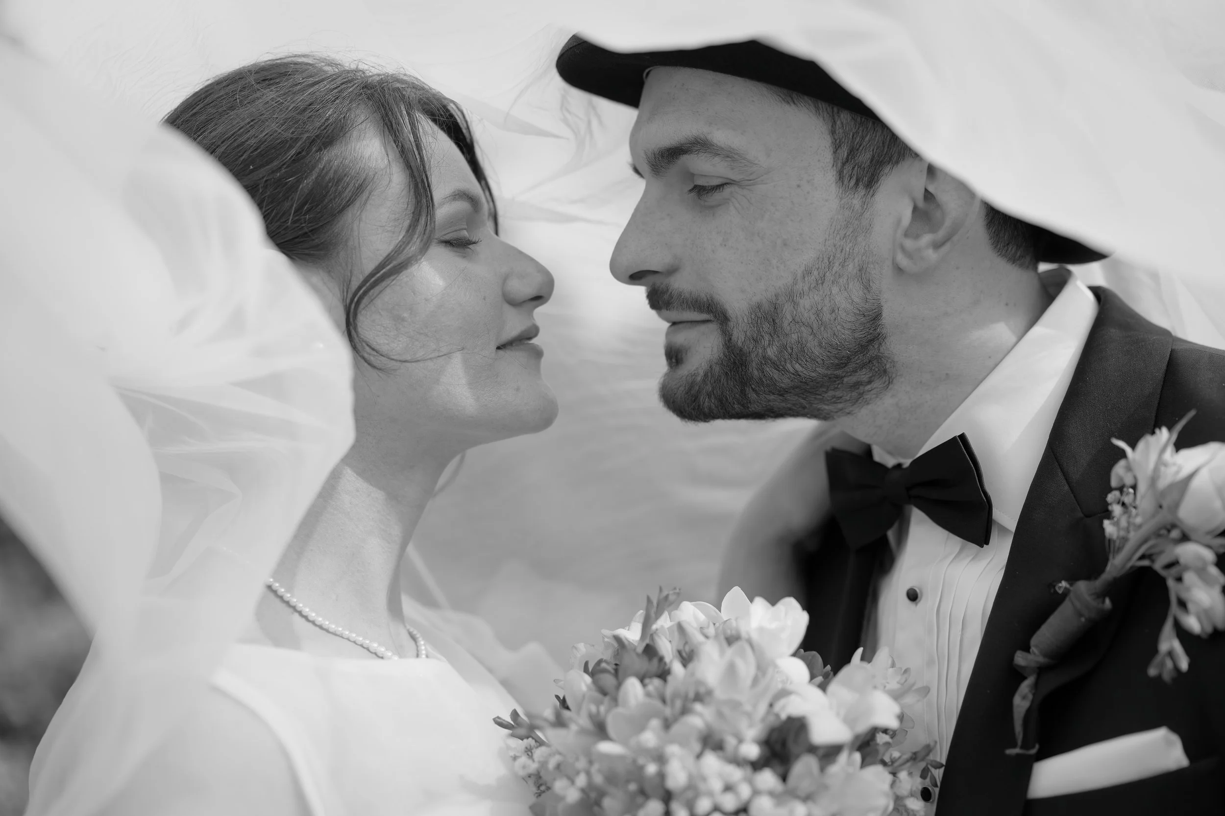 A black and white photo of a bride and groom close together, with their faces nearly touching, under a veil, on their wedding day.