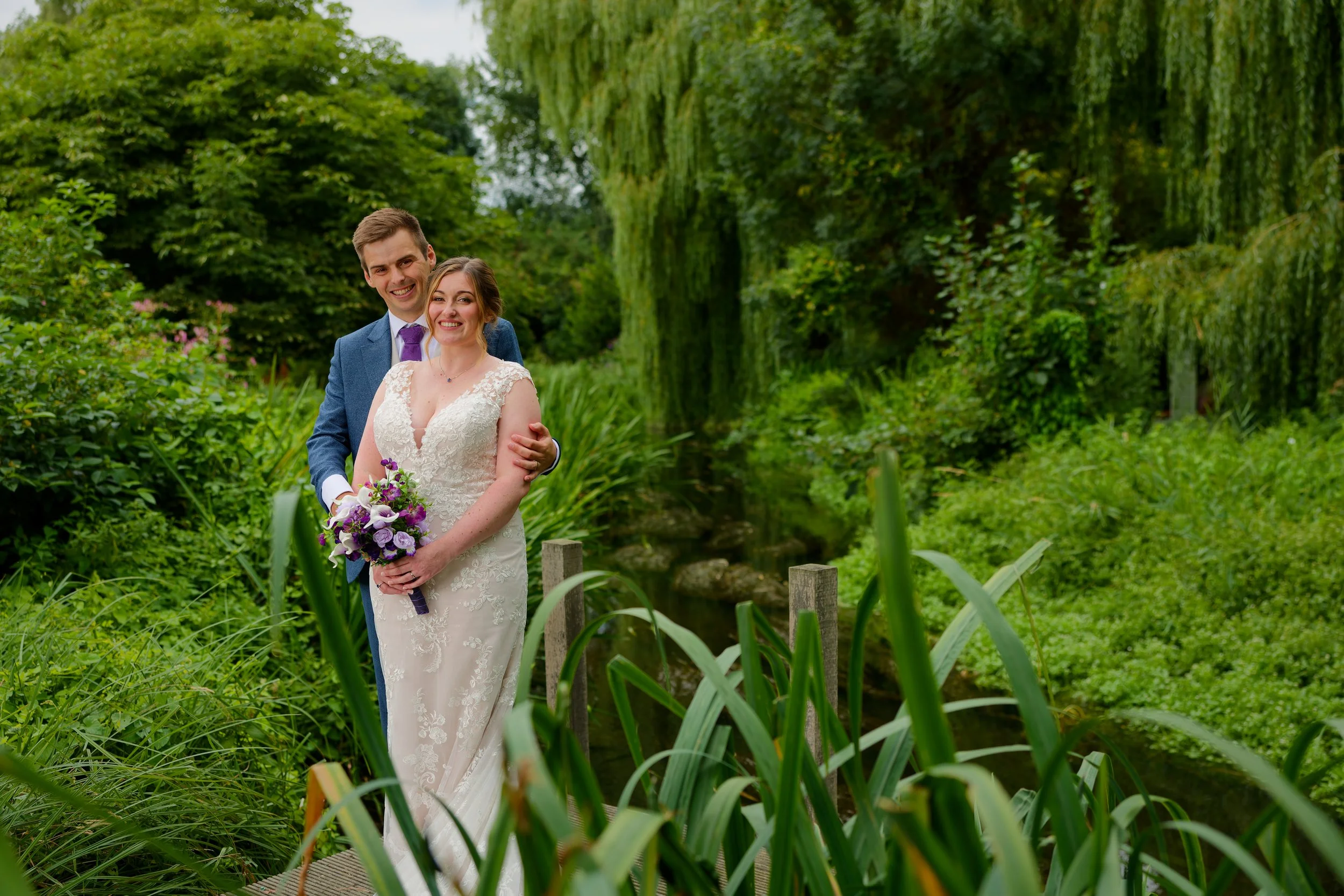 A newlywed couple standing on a small wooden bridge in a lush green garden, with the bride holding a purple and white bouquet and the groom behind her, both smiling.