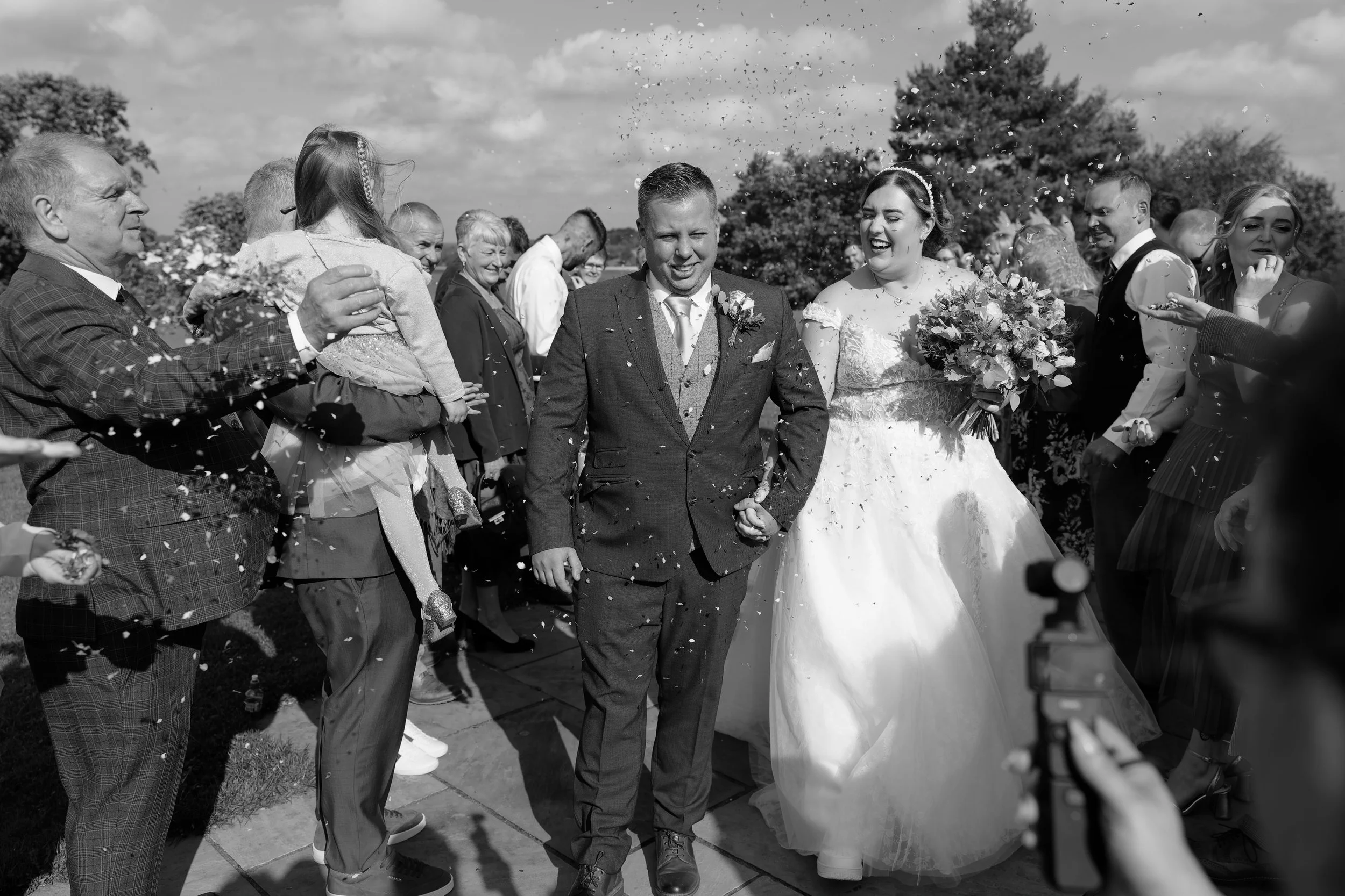 A black-and-white photo of a newlywed couple walking through a crowd of friends and family, celebrating and throwing rice or confetti at a wedding outdoors.