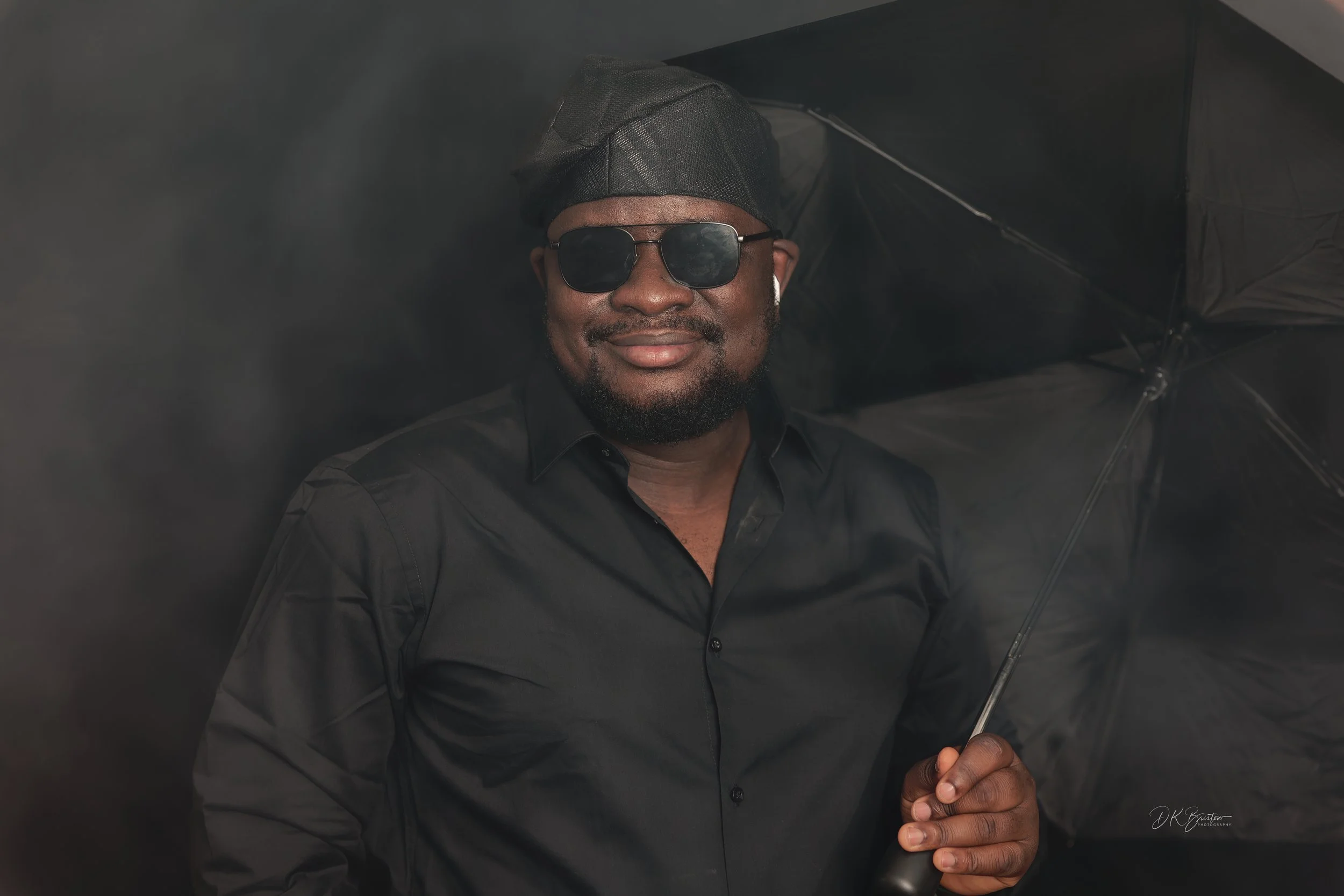 Man wearing black sunglasses, black shirt, black cap, holding black umbrella, smiling, dark background.