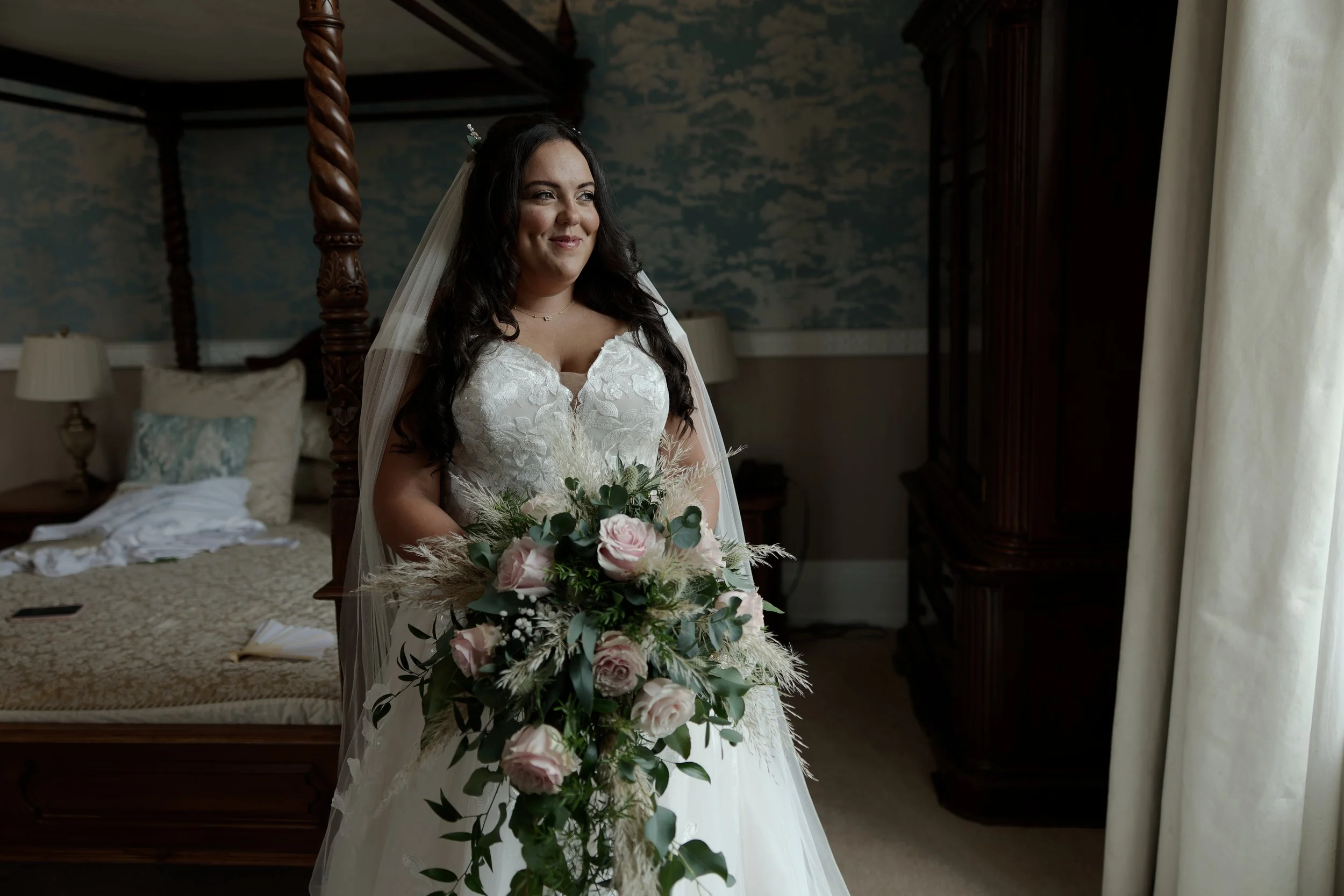 A smiling bride in a white wedding dress standing in a bedroom, holding a large bouquet of pink roses, greenery, and pampas grass, near a window with cream curtains.