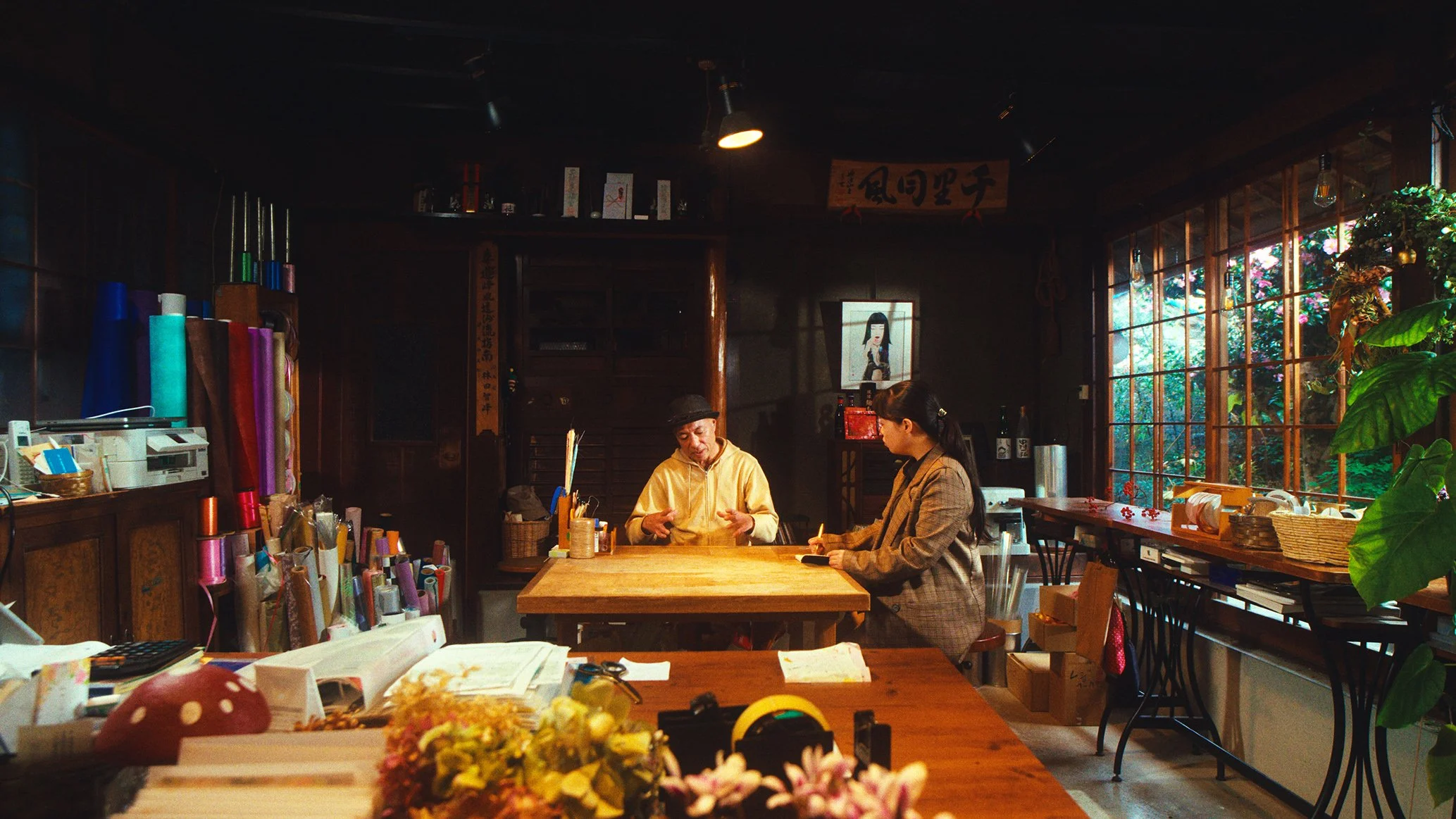 木製のテーブルに二人の女性と男性が座っている。背景には大きな窓から緑の植物が見える。