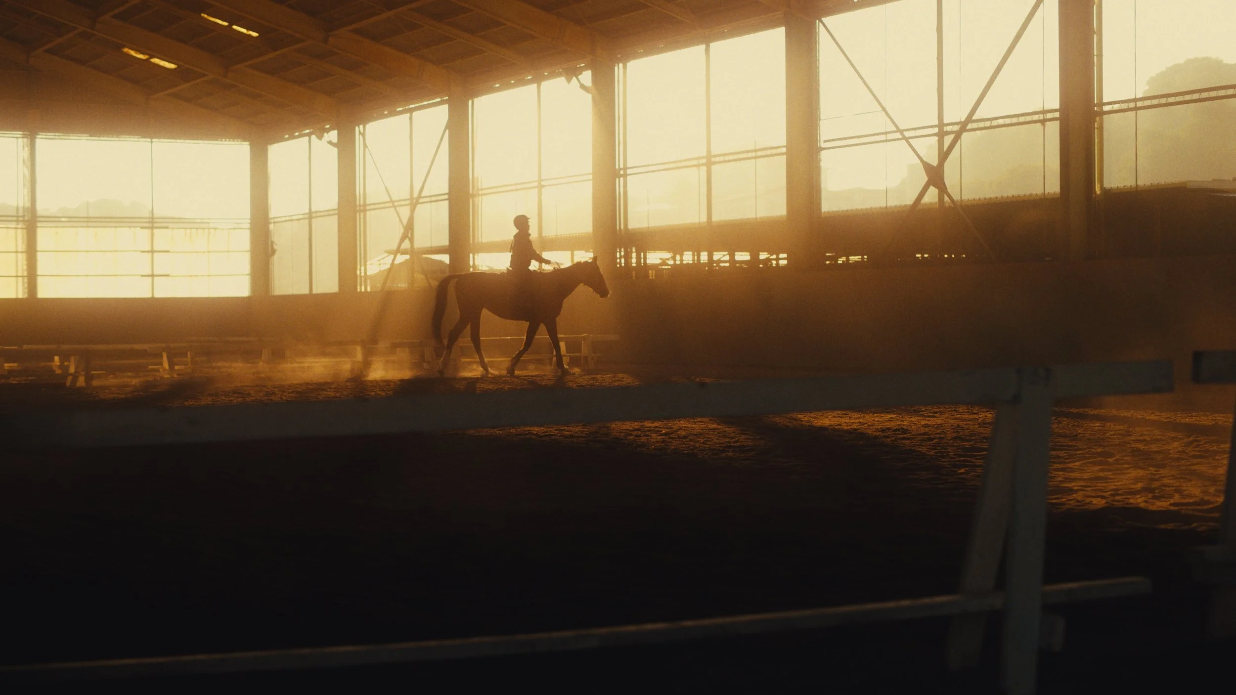 馬に乗る人が屋内の馬術練習場を馬で歩いている。夕焼けの光が差し込み、温かみのある光景。
