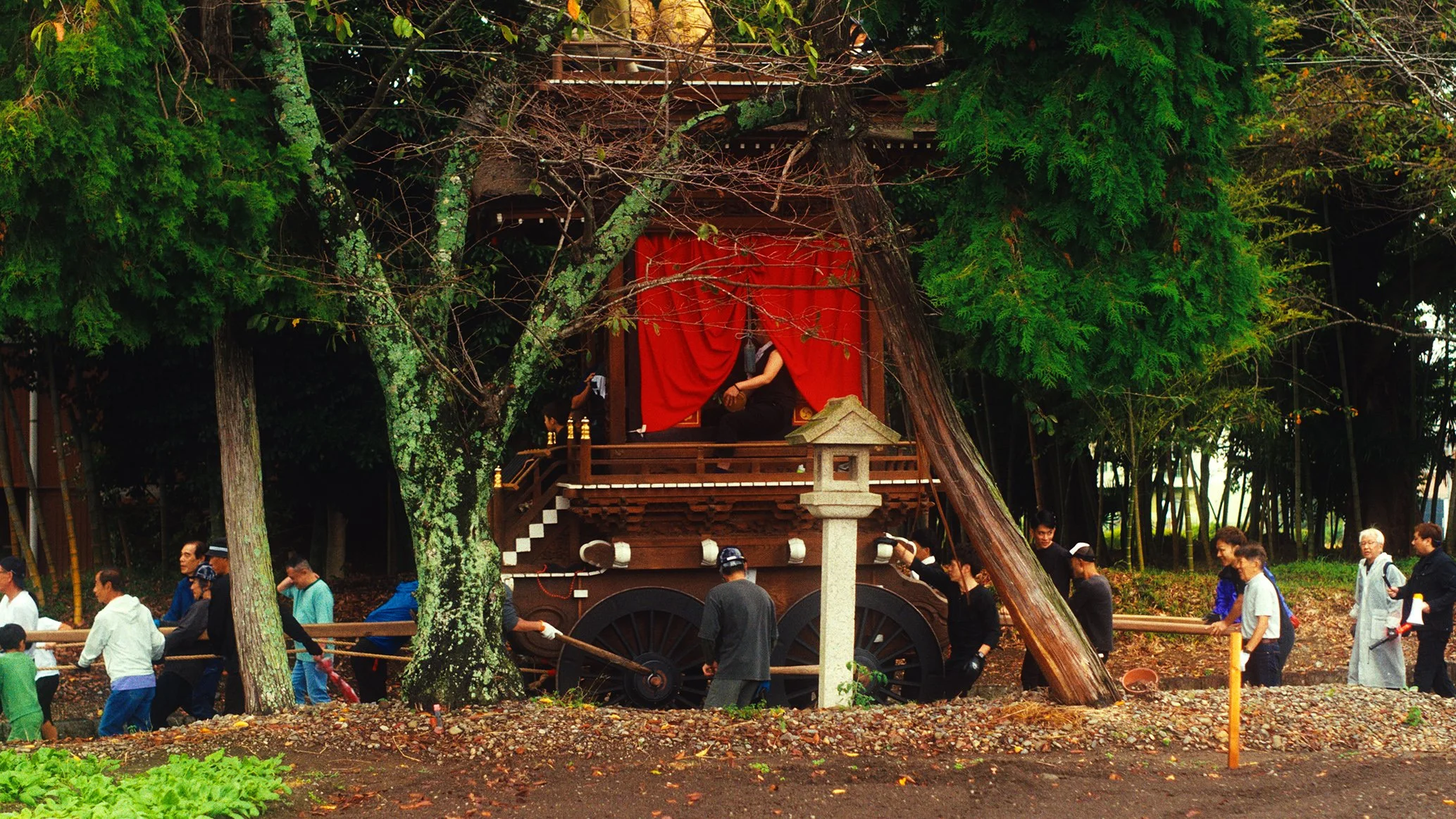神輿を引く人々と神楽舞台の風景。背景に木々が生い茂る。