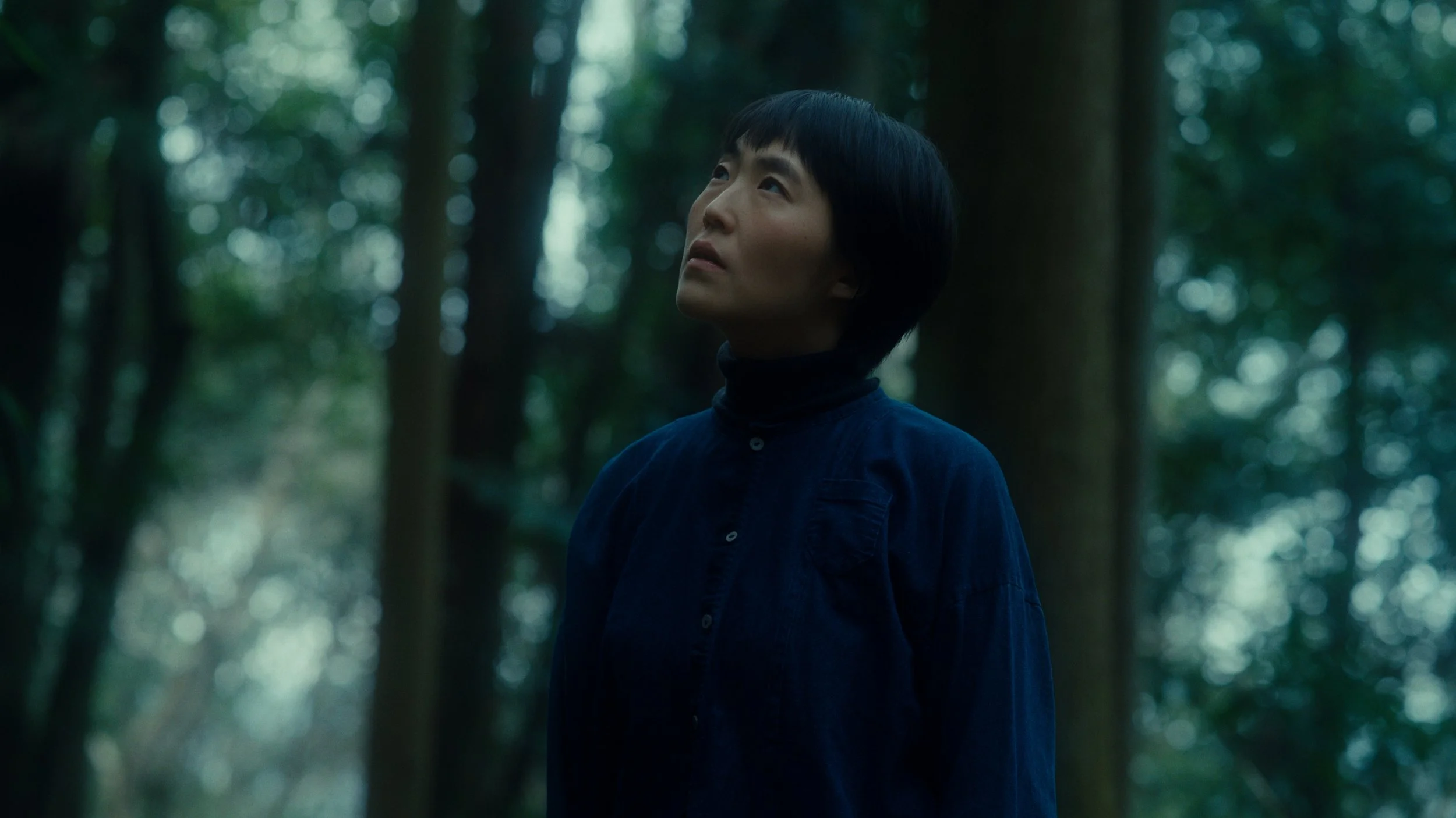 森の中で立つ女性の横顔。暗い緑色の木々に囲まれた静かな風景。