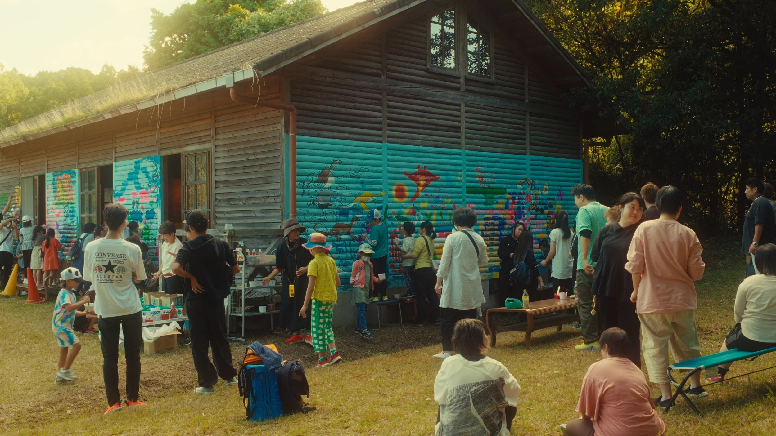木造の建物の壁に子供たちがクレヨンで描いたカラフルな絵がある屋外イベントの様子。多くの人が集まり、遊んだり見たりしている。