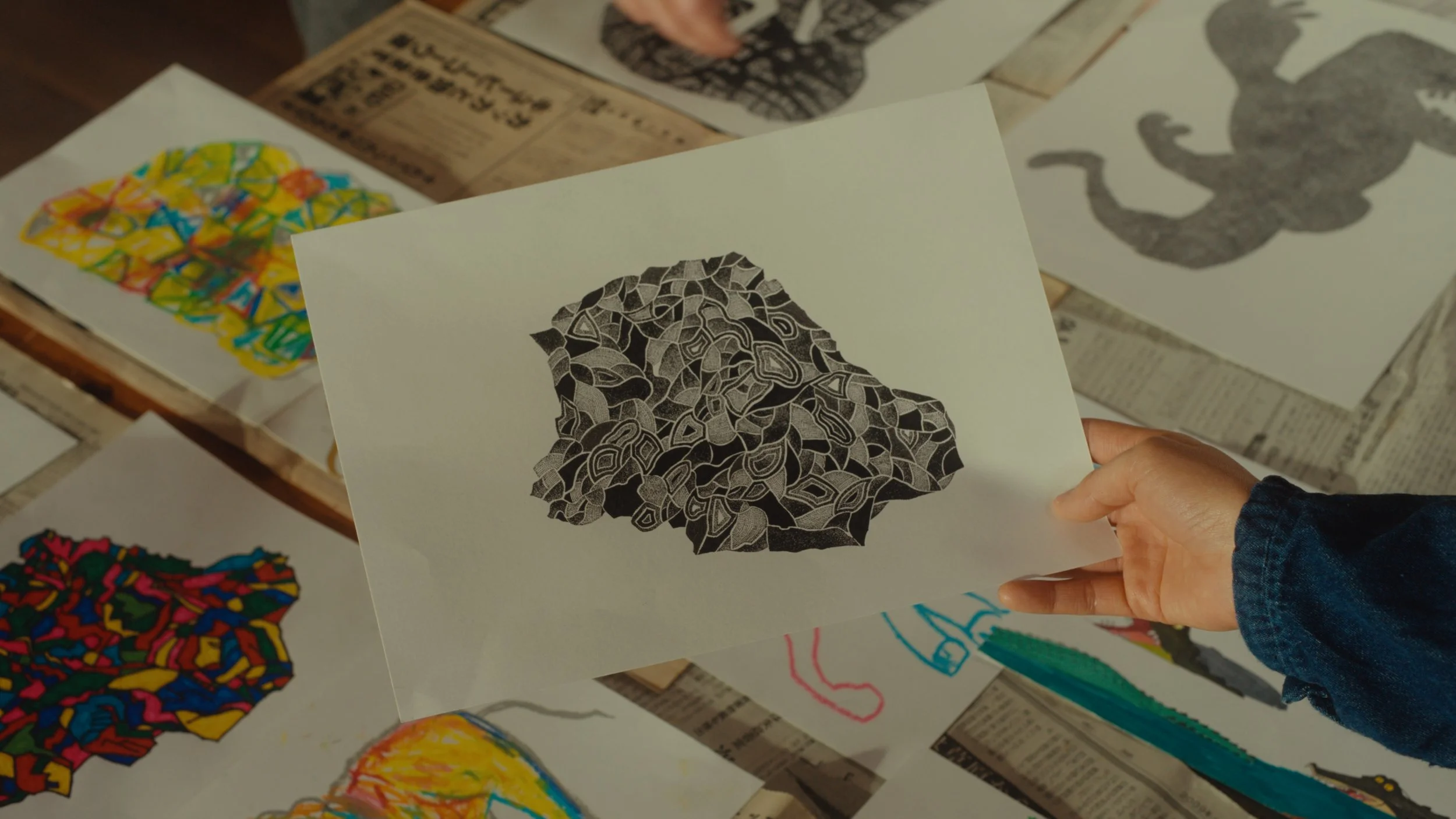 手がカラーとモノクロの抽象アート作品が描かれた紙を持っている。背景には複数の作品と新聞紙が見える。
