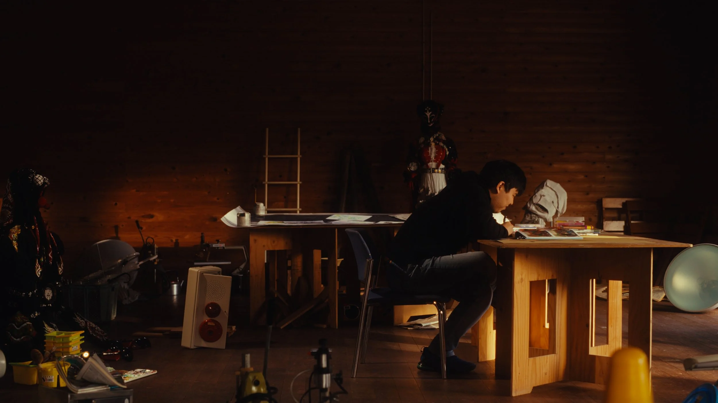 木の壁の部屋で、一人の男性が机に向かって集中している。背景にはマネキンや照明、さまざまな雑然とした物が置かれている。