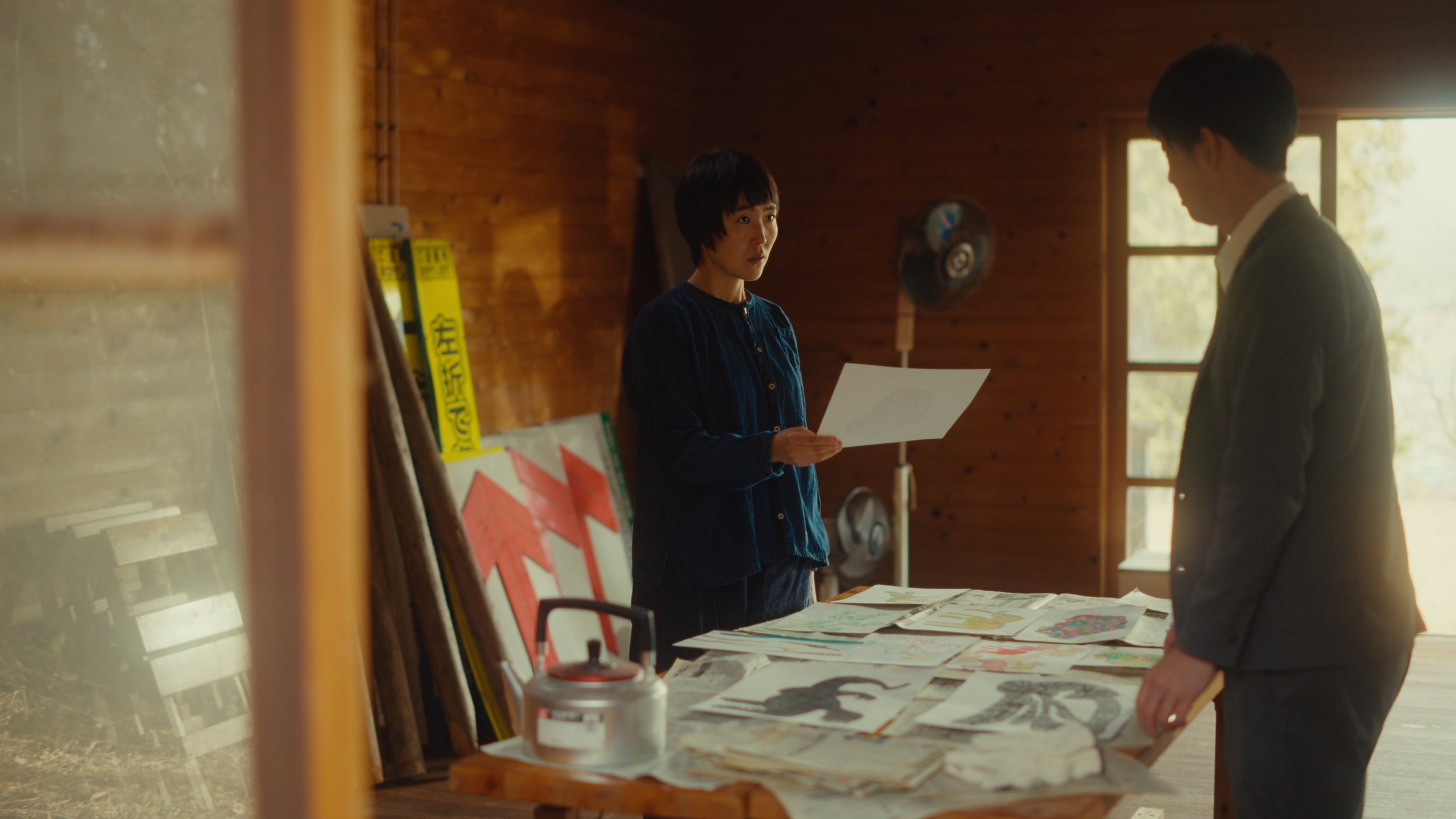 木の壁の部屋で、女性が資料を持ち男性に説明している。テーブルには新聞や雑誌が広がっている。