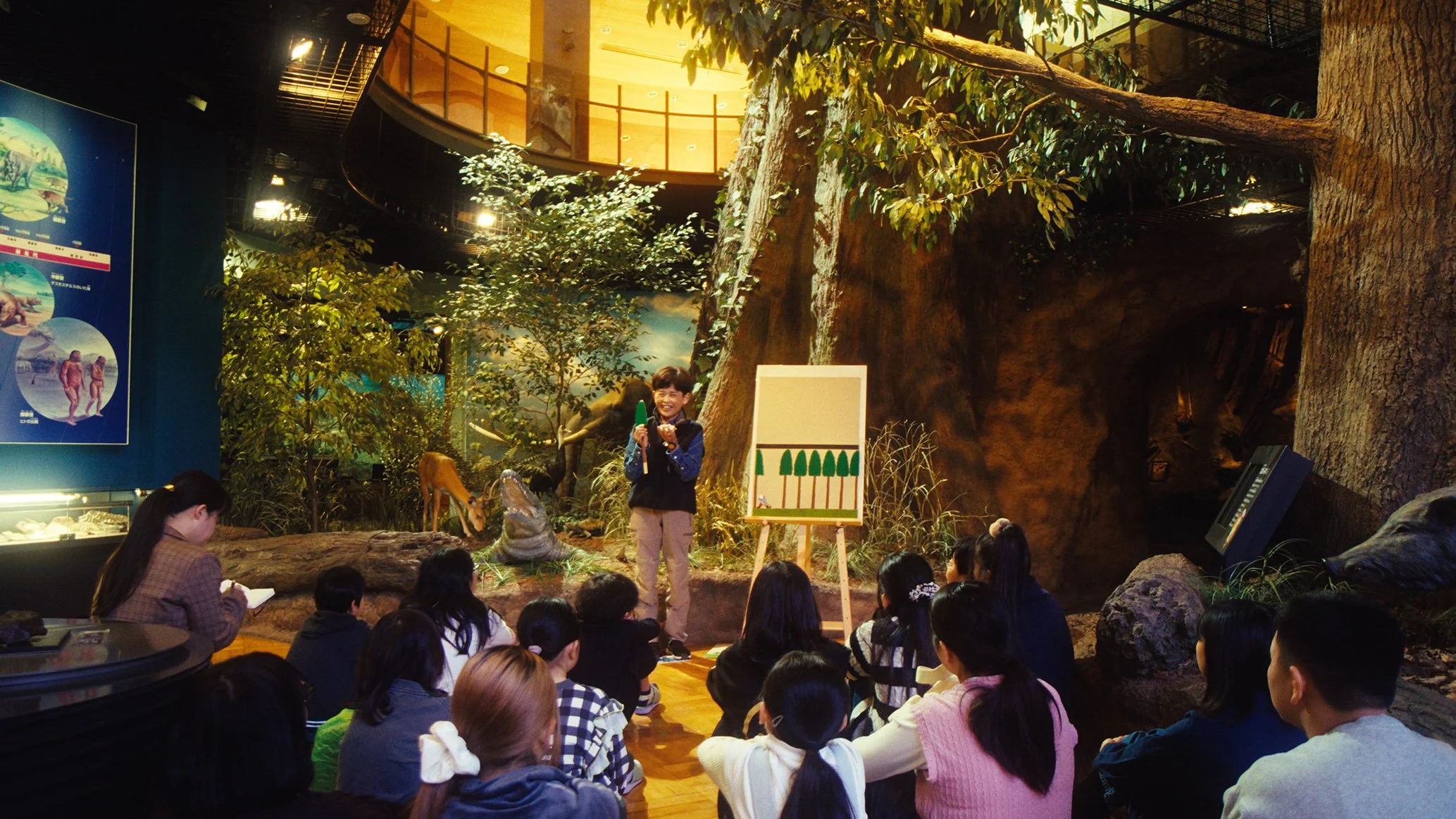 動物テーマの展示室で子供たちが恐竜や動物について学ぶためのレクチャーを受けている。講師の女性が話しながら、葉の模型を指して説明している。背景には木や恐竜の模型がある。