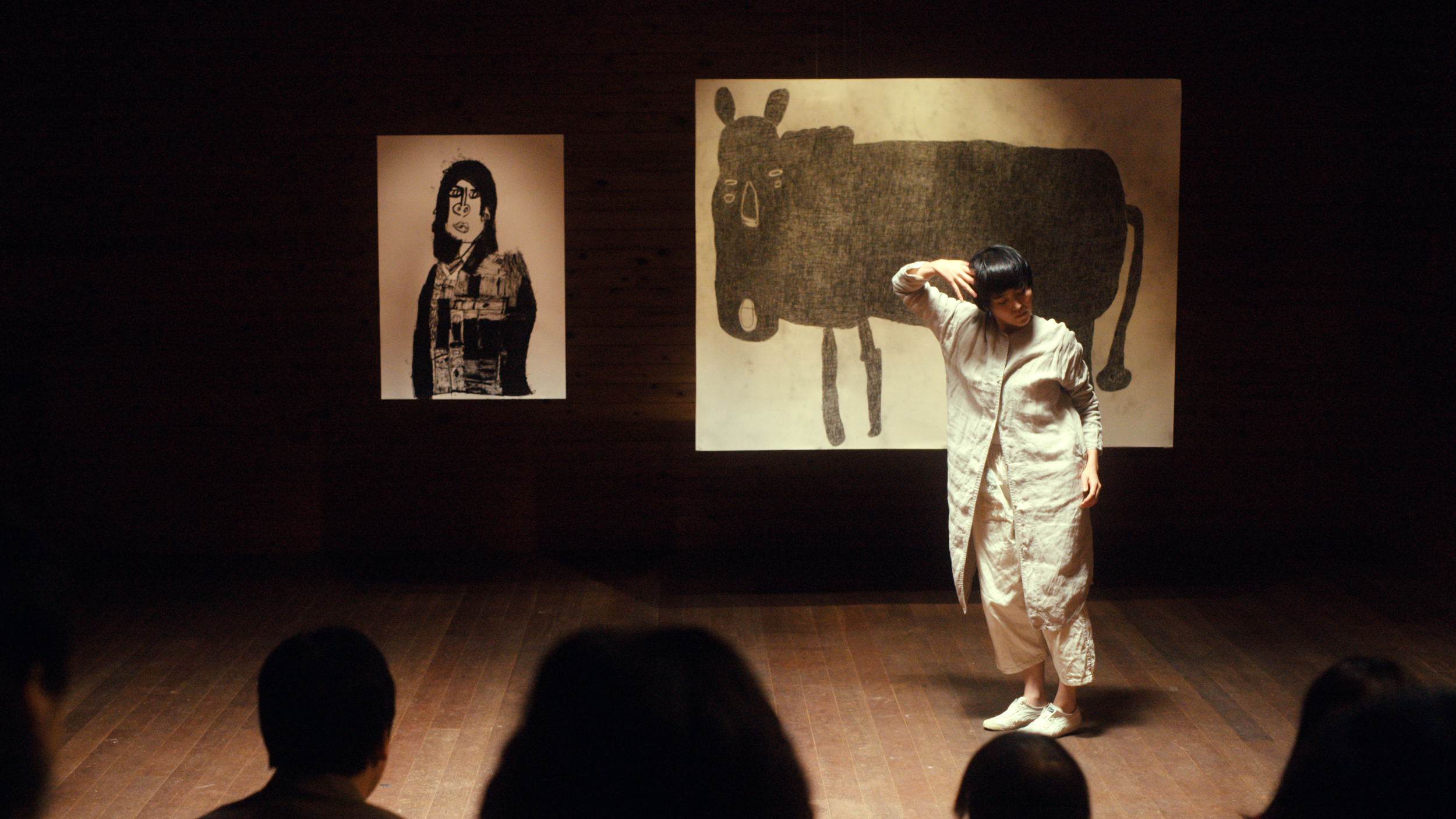 舞台でパフォーマンスをする女性と、背景に黒い動物の絵と人物の絵が飾られています。