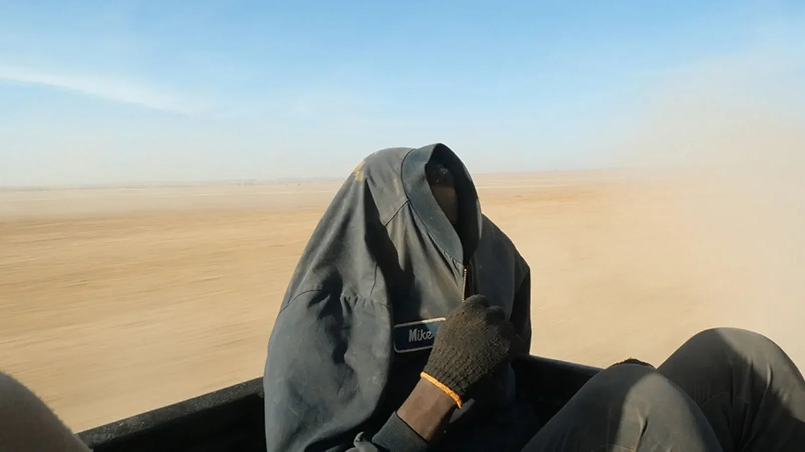 Filmstill aus ON THE BORDER: Eine vermummte Person sitzt auf der Ladefläche eines Fahrzeugs in der Sahara.